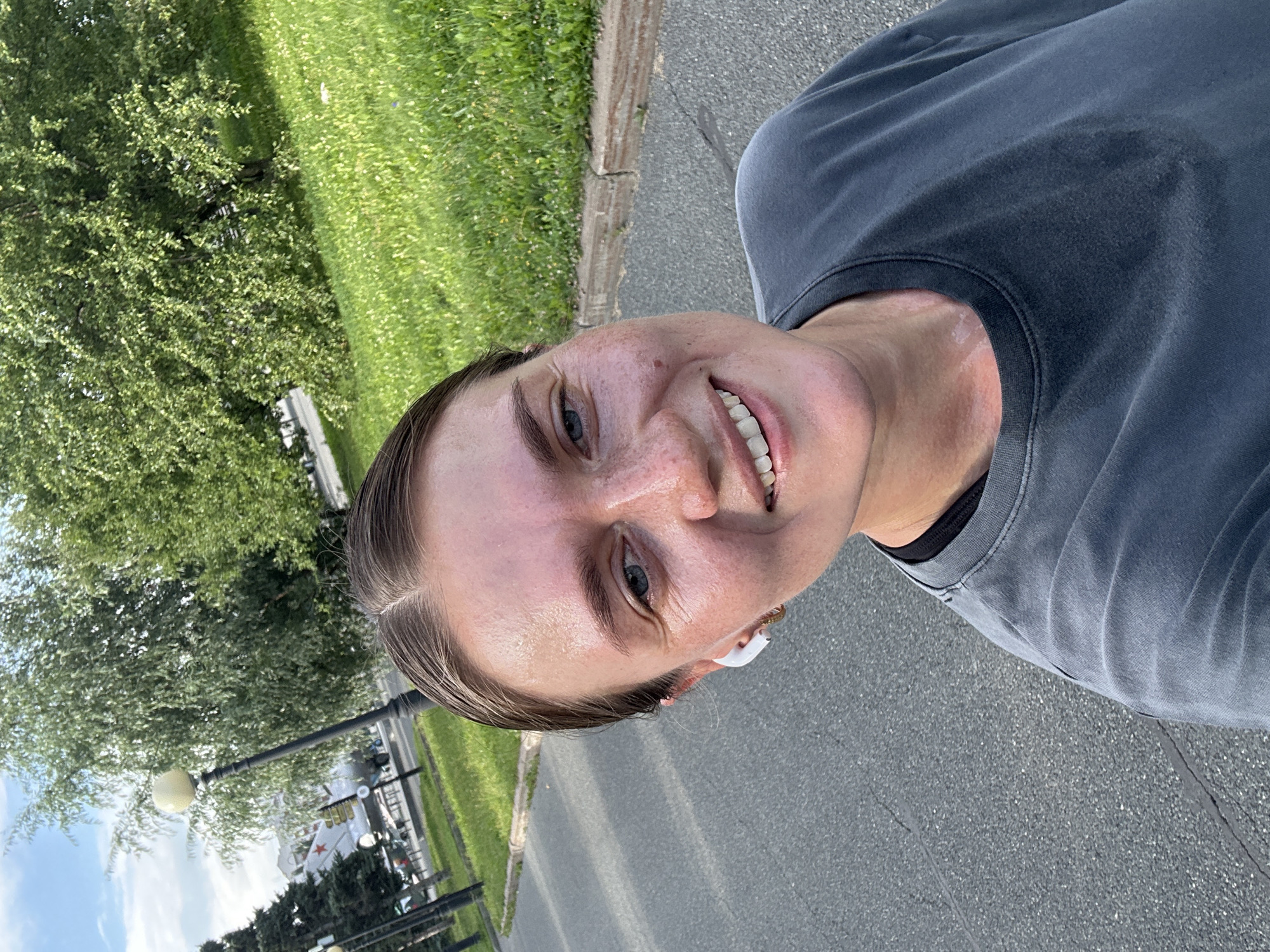 A selfie is taken by a young woman with light skin, brown hair pulled back, and blue eyes, smiling as she runs outdoors on a paved path. She is wearing a dark gray t-shirt, which appears damp, indicating she has been exercising. White wireless earbuds are visible in her ears, and a gold hoop earring is in her left ear. She has freckles across her cheeks and nose.
Behind her, a paved path runs parallel to a grassy slope dotted with small white flowers. In the background, several trees with green foliage line the path, and a streetlamp with a spherical light fixture is visible. Further back, a portion of a building and what appears to be a monument or aircraft on a pedestal can be seen. The sky is partly cloudy. The location is identified as Kazan, Russia.