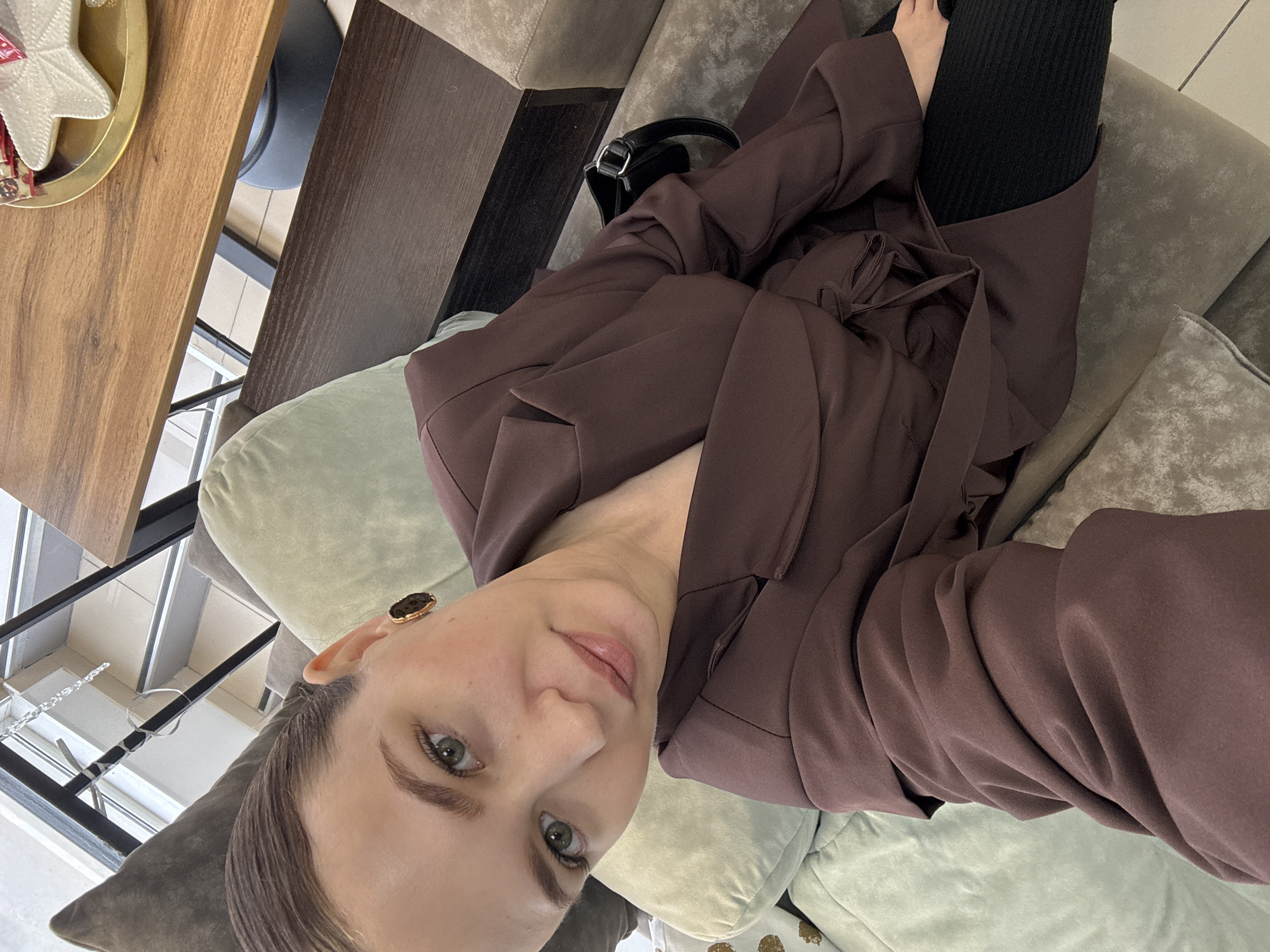 A woman with brown hair pulled back is seated on a couch, facing the camera. She is wearing a dark brown blazer over black pants and a dark accessory is visible on the couch behind her. Her left ear is adorned with a dark, square-shaped earring. The woman is looking directly at the camera with her head tilted. The background shows a blurred indoor setting with furniture. No specific Chaikovsky, Russia landmarks or context are discernible in the image.