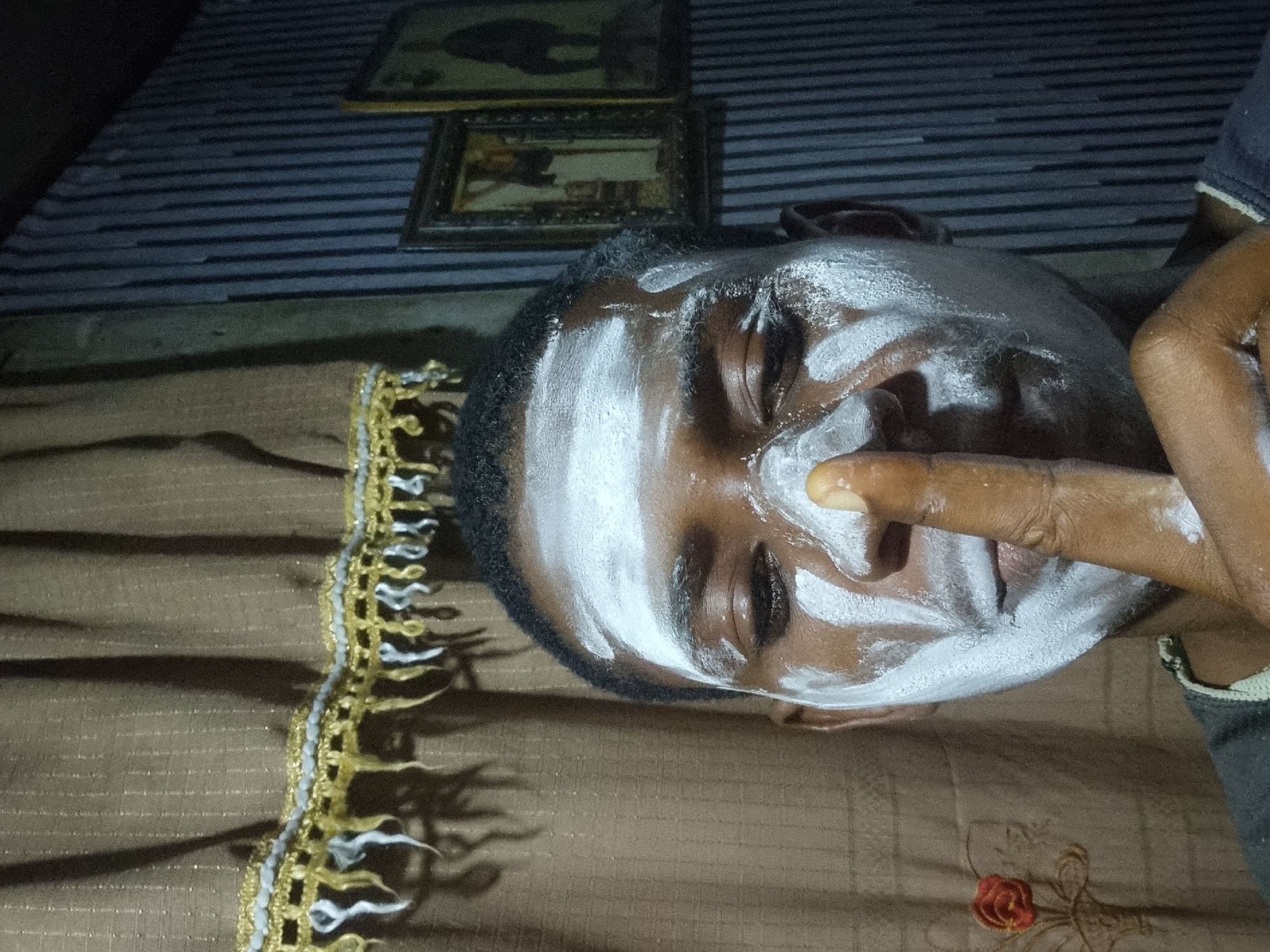 An African man lies on a brown blanket, his face covered in white cream. His right hand is near his face, the index finger pointing up and covered in the cream. The background features a striped surface and two framed pictures. The location is Onitsha, Nigeria.