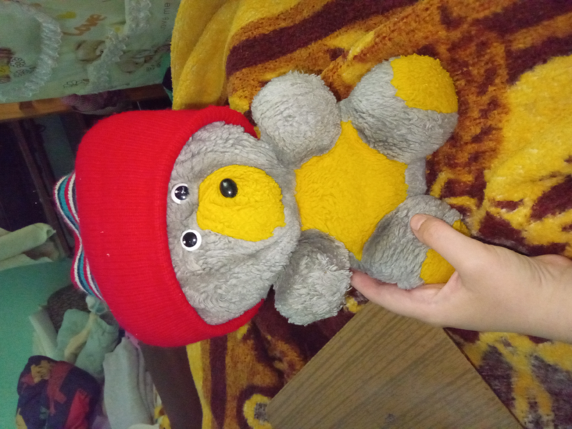 A stuffed teddy bear with gray fur and a yellow belly and feet wears a red knitted hat with a striped brim. The bear is sitting on a brown and yellow striped blanket. A human hand is visible holding the bear's left arm. In the background, there are various soft objects, possibly bedding or clothing, and a wooden surface. The scene appears to be indoors, in a domestic setting.