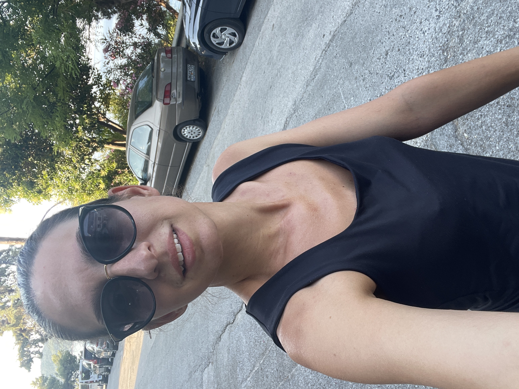 A woman with dark hair pulled back and wearing large, black-rimmed sunglasses, a black tank top, and a gold necklace stands on a paved street in Sithonia Municipal Unit, Greece. She is holding a camera, presumably taking a selfie. In the background, several cars are parked along the street, and lush green trees and foliage are visible. The sunlight casts shadows, indicating it is daytime. A tan car with a license plate reading "HMH1420G" is prominent in the mid-ground. The overall setting appears to be a residential or tourist area.