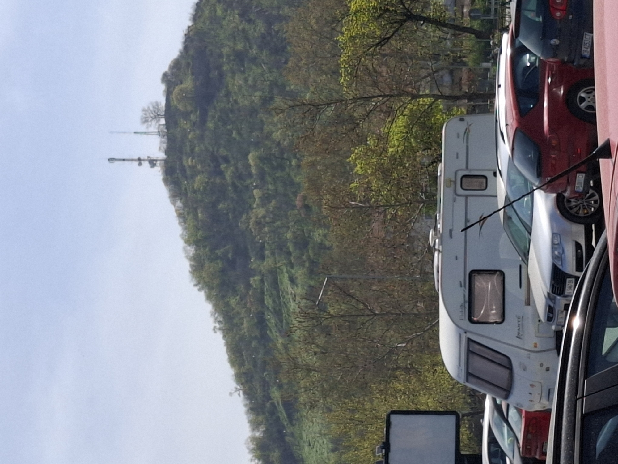 This image, rotated 90 degrees counter-clockwise from its likely original orientation, depicts a serene daytime scene in Plovdiv, Bulgaria, featuring parked vehicles against the backdrop of a large, tree-covered hill.
In the foreground, a row of vehicles is parked along what appears to be a street or parking area. From left to right (if the image were rotated correctly): a partially visible black car; a prominent white caravan with the text "AVANTE" on its side, featuring a large front window and a smaller side window; a silver/grey car parked behind the caravan; a red hatchback or sedan; and a dark blue Volkswagen Golf Mk5, whose Bulgarian license plate (P 3425 JC) is partially visible. A black antenna-like object extends into the bottom right of the frame, possibly from an unseen vehicle, and a white rectangular sign or object is partially visible in the bottom left.
The middle ground is dominated by a lush, green hill, heavily forested with deciduous trees, suggesting spring or summer. At the summit of the hill, two distinct communication towers or masts stand tall, accompanied by a bare-branched tree.
The sky in the background is clear and bright blue, indicating sunny weather. No specific human activity is visible, making the scene appear still and quiet. The distinct hill and the Bulgarian license plate are strong location cues for Plovdiv.