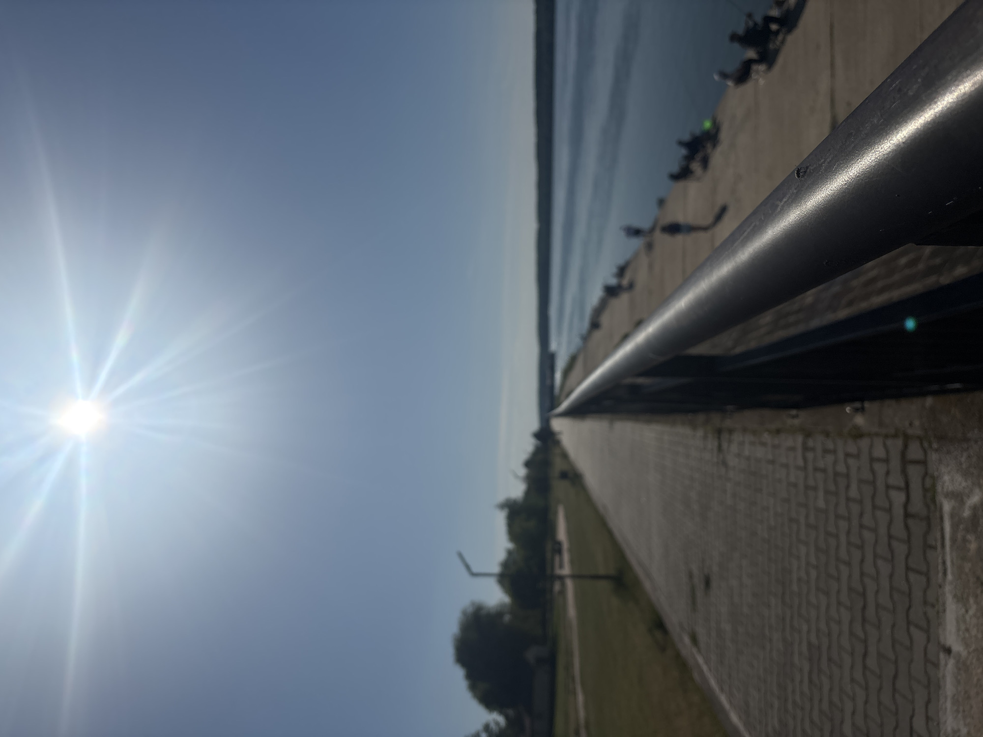 This image captures a scene along the Danube River in Lom, Bulgaria. The photo is taken from a low angle, possibly near a railing. A paved walkway made of interlocking gray bricks stretches into the distance, bordered by a grassy area with scattered trees and light posts. The Danube River appears to the left, with the opposite bank visible in the distance. Silhouetted figures are visible along the river's edge on what appears to be a concrete bank. The sky is bright, with the sun's glare dominating the left side of the frame. A metal railing runs horizontally across the upper right portion of the image.