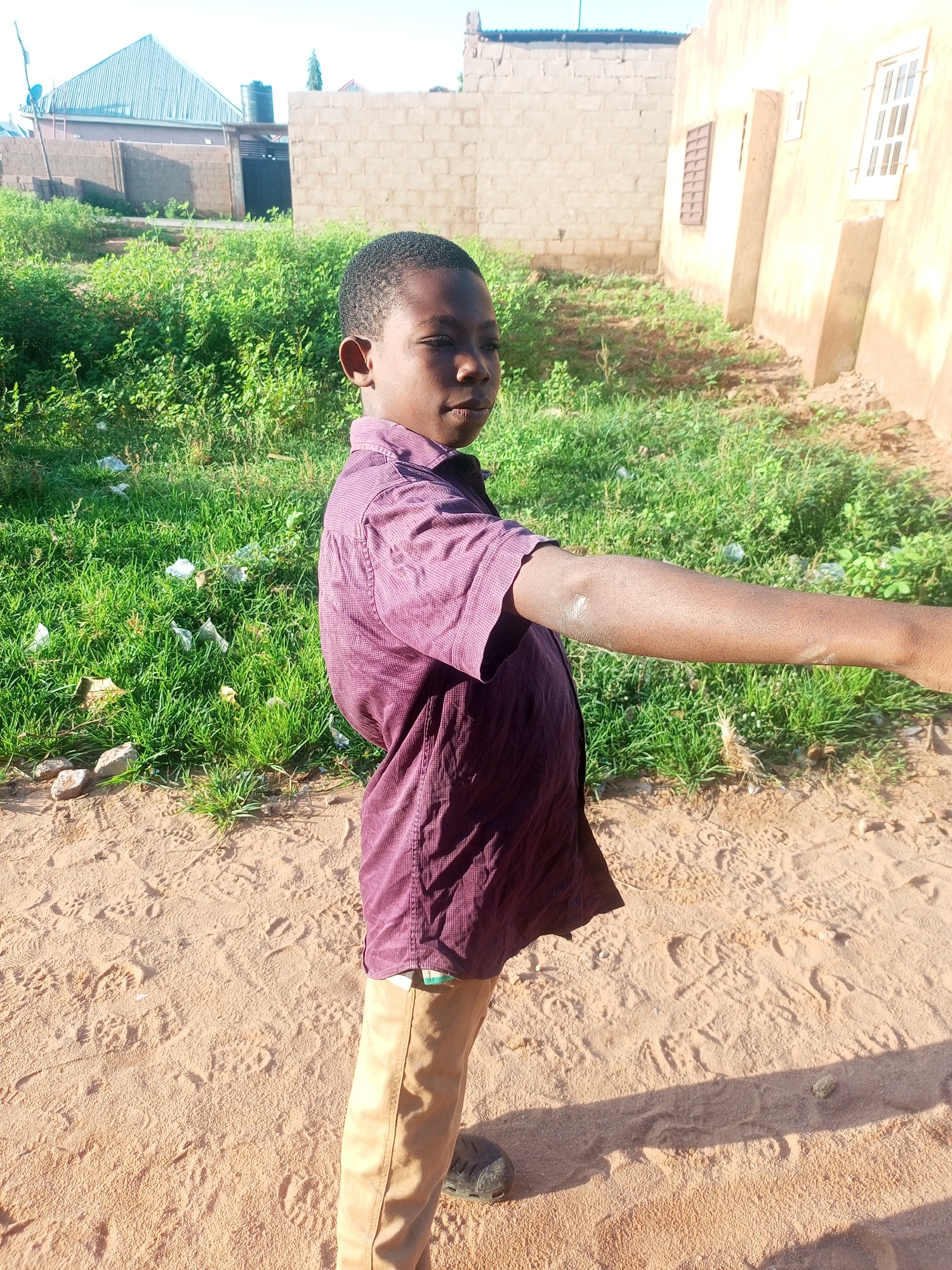 The image depicts a young boy standing on sandy ground in what appears to be a rural or semi-urban area. The boy is wearing a dark purple collared shirt and tan pants. He is looking to his right, with his right arm extended outwards. The background consists of green vegetation, some scattered debris, and buildings constructed of brick and concrete. One building in the background has a light yellow facade and windows with metal bars. Another building has a corrugated metal roof. The sky is clear and bright. The environment suggests an outdoor setting in a warm climate. The presence of the buildings and the sandy terrain points to a location within Tempure, Nigeria, characterized by its typical architectural styles and natural landscape.