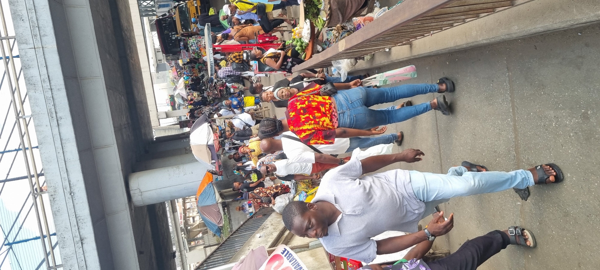 The image shows a bustling market scene in Ikeja, Nigeria, situated underneath a concrete overpass. Numerous vendors and shoppers are present, creating a crowded environment. Merchandise such as fruits, vegetables, and general goods are displayed on makeshift stalls or directly on the ground. People are dressed in a variety of casual clothing. The ground appears to be paved and slightly worn. The overpass provides overhead cover, with metal railings visible along the edge. A yellow commercial bus is partially visible in the background.