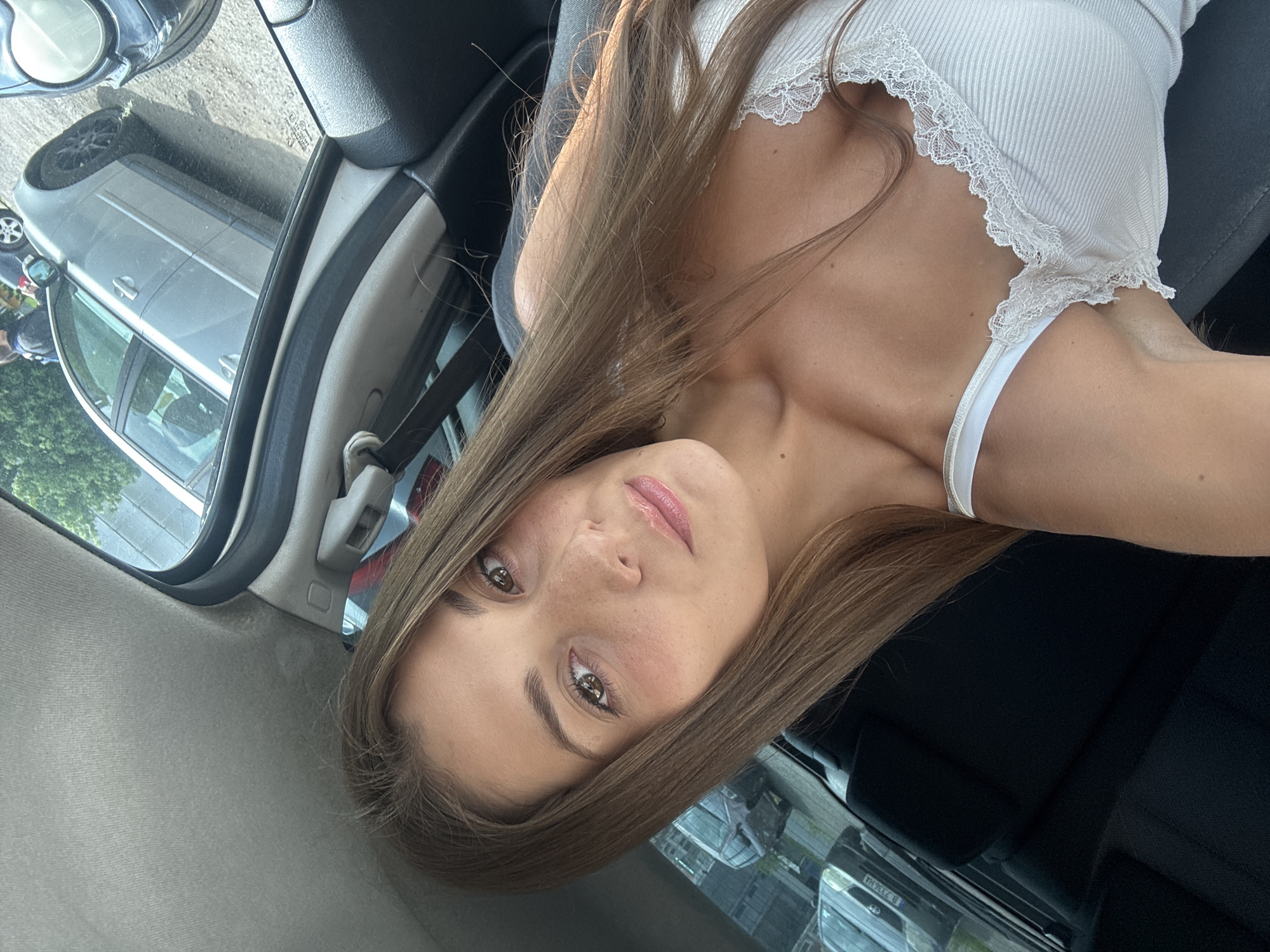A young woman is seated inside a car, presumably a passenger. She has long brown hair and is wearing a white top with lace trim. The view through the car window shows other parked cars and buildings, suggesting an urban setting. The specific location in Burgas, Bulgaria, cannot be determined from the given image.
