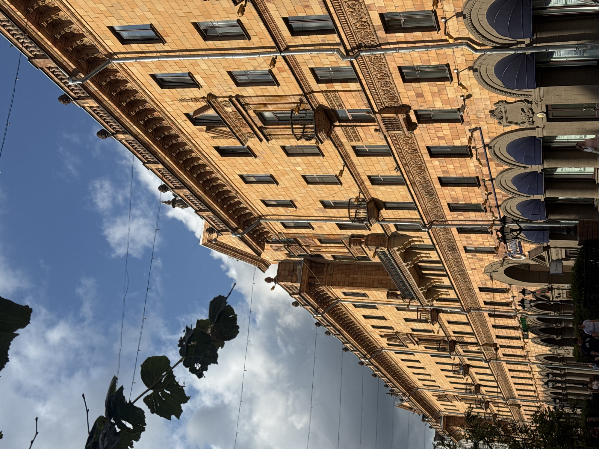 The image shows a long, ornate building facade in Minsk, Belarus, likely on Independence Avenue, a prominent street. The building is constructed with warm, orange-toned brick and features classical architectural elements such as arched windows, decorative cornices, balconies, and sculptures. The sky above is blue with scattered white clouds, and some dark green foliage from trees is visible in the upper left corner. Several strings of small lights are strung across the sky in front of the building. The lower portion of the building features a row of arched storefronts with blue awnings. A few people are walking on the sidewalk in front of the building, and some are visible through the store windows. The overall impression is of a grand, historic urban environment.
