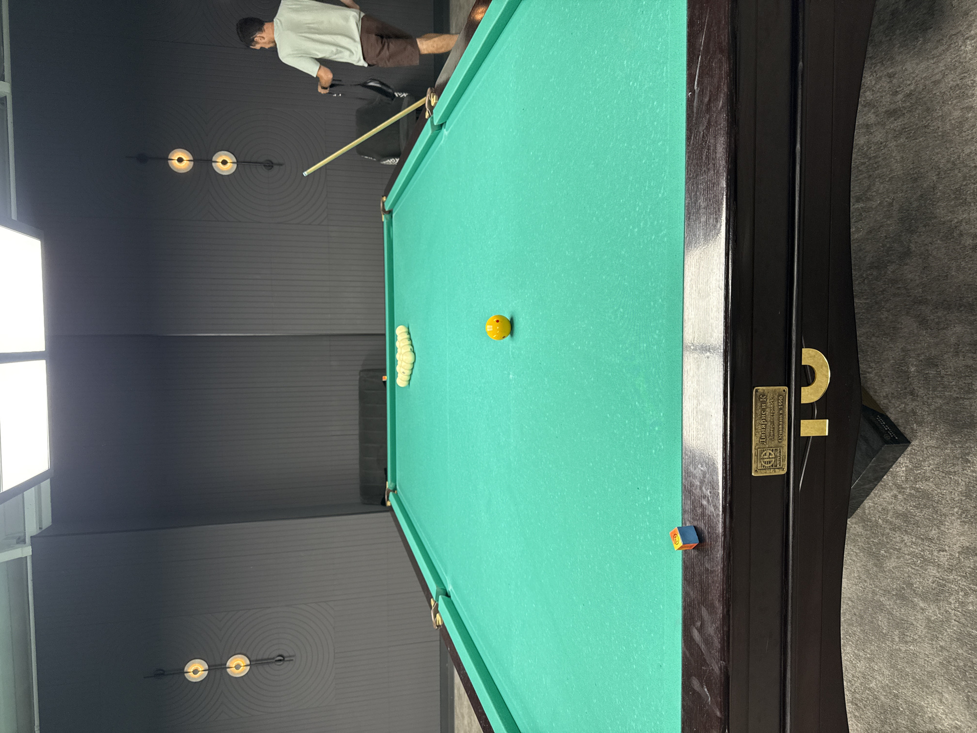 The image shows an indoor billiards room with a green felt pool table in the foreground. The table has a dark wooden frame with brass accents. A yellow billiard ball is in the center of the table, and a rack of white balls is positioned at one end. A small, multi-colored cube is on the edge of the table. A man in a light-colored t-shirt and brown shorts stands near the table, holding a cue stick and a strap. The room has dark gray walls with decorative circular patterns and two sets of wall-mounted lights. The floor appears to be carpeted. The scene is consistent with a billiards hall or recreational area in a city like Balaklavsky District, Ukraine.