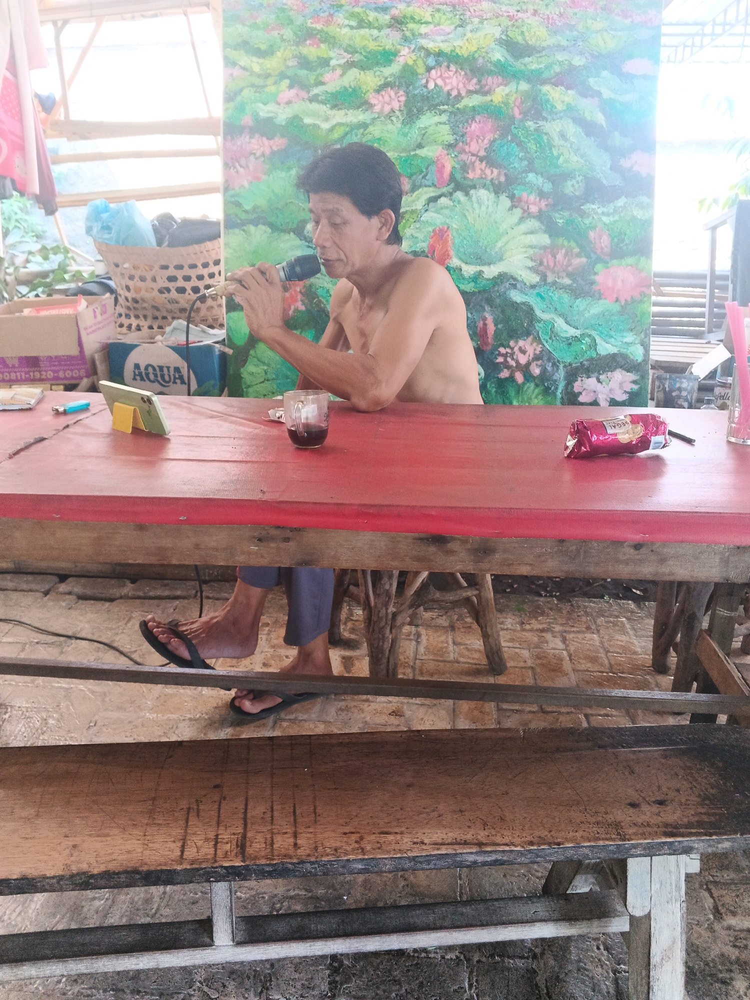 An Indonesian man is sitting at a red table and speaking into a microphone. He is shirtless and wearing blue pants and flip-flops. In front of him on the table is a glass of dark liquid, a smartphone on a stand, a lighter, and a packaged snack. Behind him, a large, colorful painting of lotuses hangs on the wall. The scene appears to be outdoors, possibly in a market or roadside stall, given the rustic wooden furniture and the natural lighting. The man's activity suggests he might be singing or giving a speech. The overall impression is one of a casual, possibly informal gathering or performance in an open-air setting within Ungaran, Indonesia.