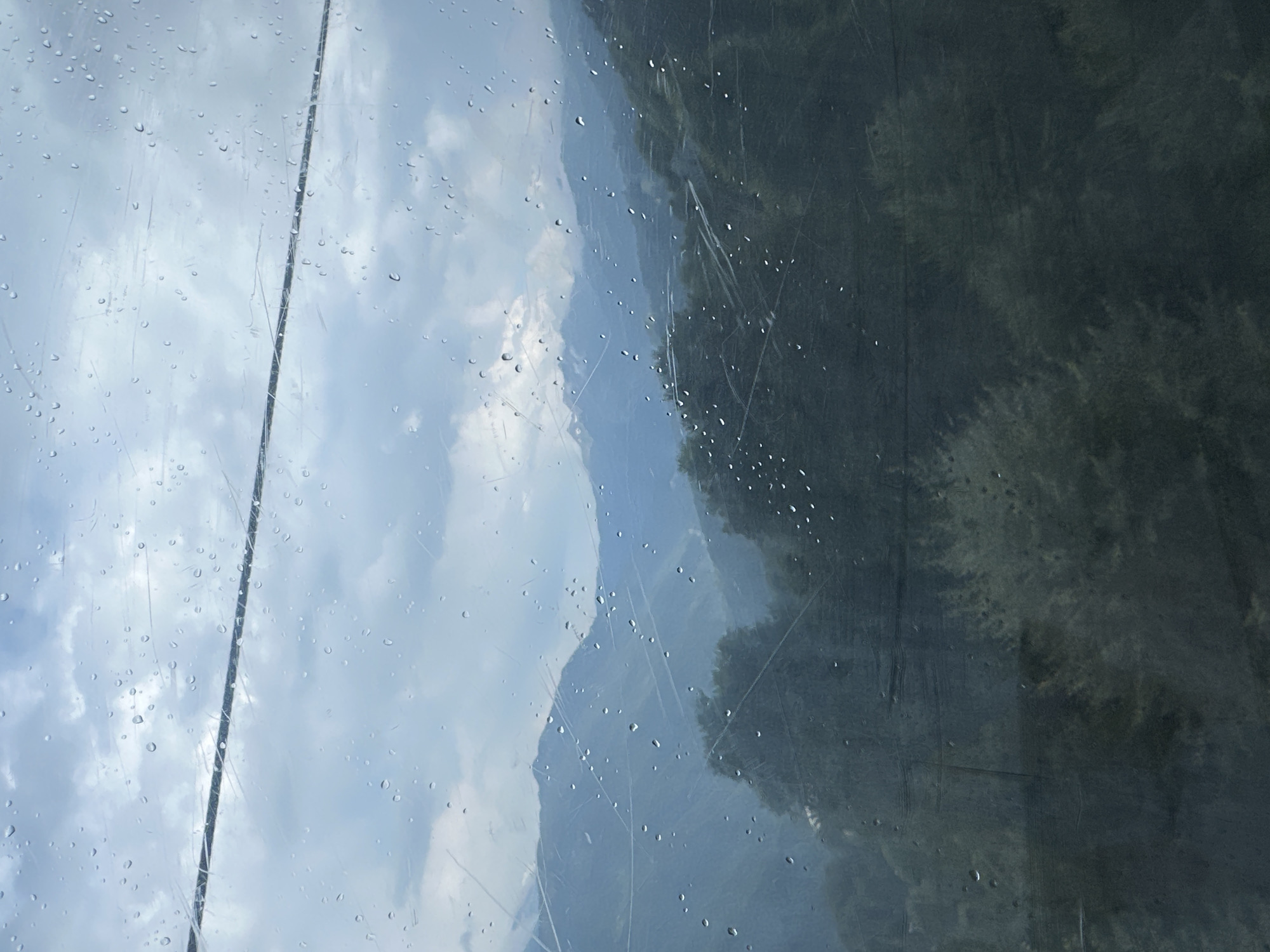 A scenic view of the mountains near Estosadok, Russia, is seen through a window streaked with raindrops. A dark cable traverses the upper part of the frame. The sky is overcast with clouds. Layers of mountains recede into the distance, with a darker, forested slope dominating the middle ground. The foreground is heavily obscured by the window's surface, with numerous water droplets and scratches distorting the view of dark vegetation. No people or specific actions are depicted.