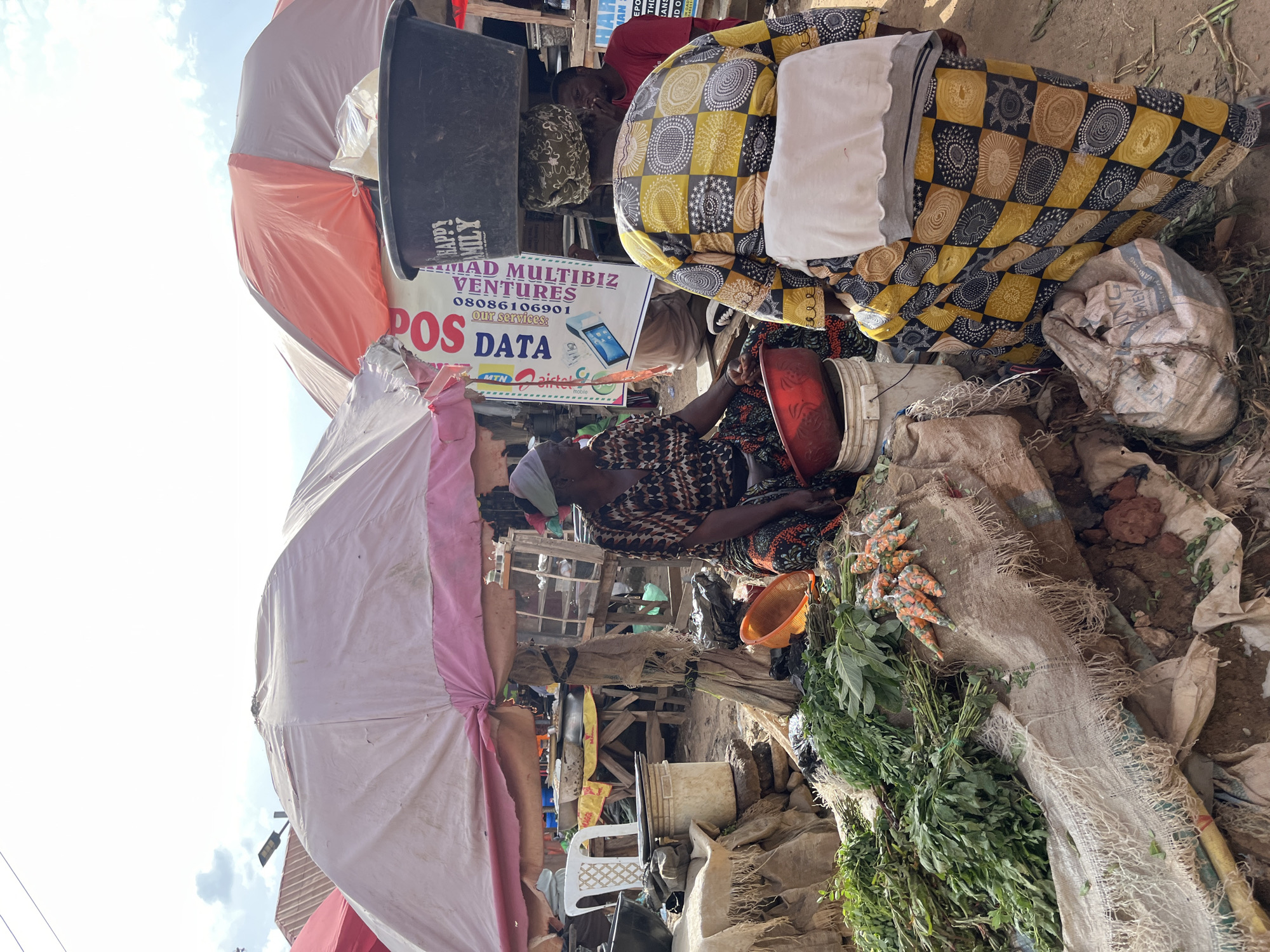 This image captures a street market scene in Rafin Par, Nigeria, under a bright sky with scattered clouds. A woman in a patterned dress is seated under a large, light-colored umbrella, surrounded by baskets and produce. To her right, another woman, wearing a vibrant yellow and black patterned outfit and a headscarf, carries a large black basin on her head. She is walking away from the viewer. In the background, a banner with text, including "POS DATA" and "HAPPY FAMILY," is visible. The ground is dusty and uneven, with various bags, baskets, and bunches of green vegetables and carrots displayed for sale. A white plastic chair is visible to the left of the seated woman. The overall impression is one of a bustling, outdoor marketplace.