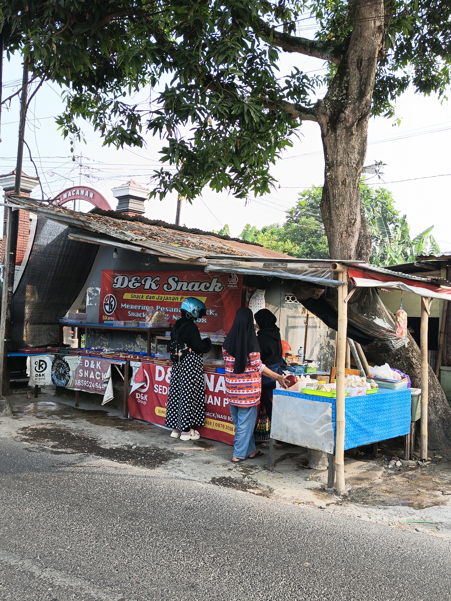 A street-level, eye-level, wide shot captures an outdoor food stall in Ketandan, Indonesia. A large tree with full green leaves shades the scene. The stall features a corrugated metal roof supported by bamboo poles. A red banner with white text, "D&K Snack," is prominently displayed, advertising snacks and accepting orders. Below the banner, various food items are displayed on shelves, including packaged snacks and what appear to be cooked goods.

Three individuals are present. Two women are seen from the back, wearing hijabs. One woman, wearing a black top and a patterned polka-dot skirt, stands near the stall, possibly interacting with a vendor. The second woman, to her left, wears a black hijab and a colorful patterned top with blue pants. A third individual, wearing a blue helmet and a black outfit with a polka-dot skirt, stands closer to the stall, looking towards the displayed food.

In front of the stall, a table is set up with a blue patterned tablecloth, displaying various food containers and packaged items, possibly prepared food for sale. A large tree trunk is visible to the right of the stall, its branches extending over the structure. The ground in front of the stall is paved and appears wet in places. The background reveals a brick building with a red arched entrance, also with the text "M. MACAN" visible. Electrical wires are strung across the scene, typical of an urban environment. The sky is overcast.
