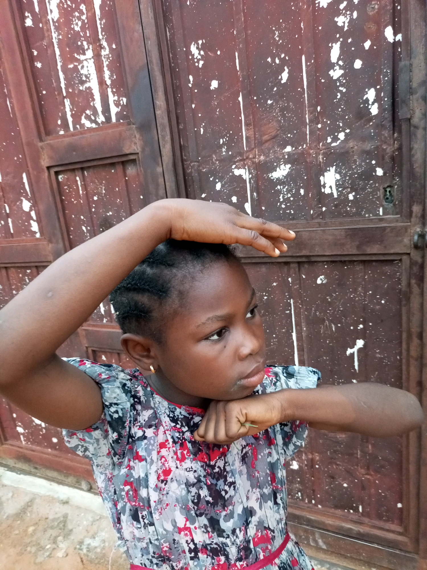 A young Black girl with dark braided hair and a patterned dress poses in front of a weathered brown door. Her left arm is raised and bent at the elbow, with her hand resting on top of her head, while her right arm is bent across her chest with her fist near her chin. She is looking to her right with a neutral expression. The door is made of vertical wooden planks and is heavily chipped and stained with white paint splatters. The ground in front of the door appears to be dirt and gravel. The setting is outdoors, possibly a residential area in Tempure, Nigeria, given the style of the door and the girl's appearance.