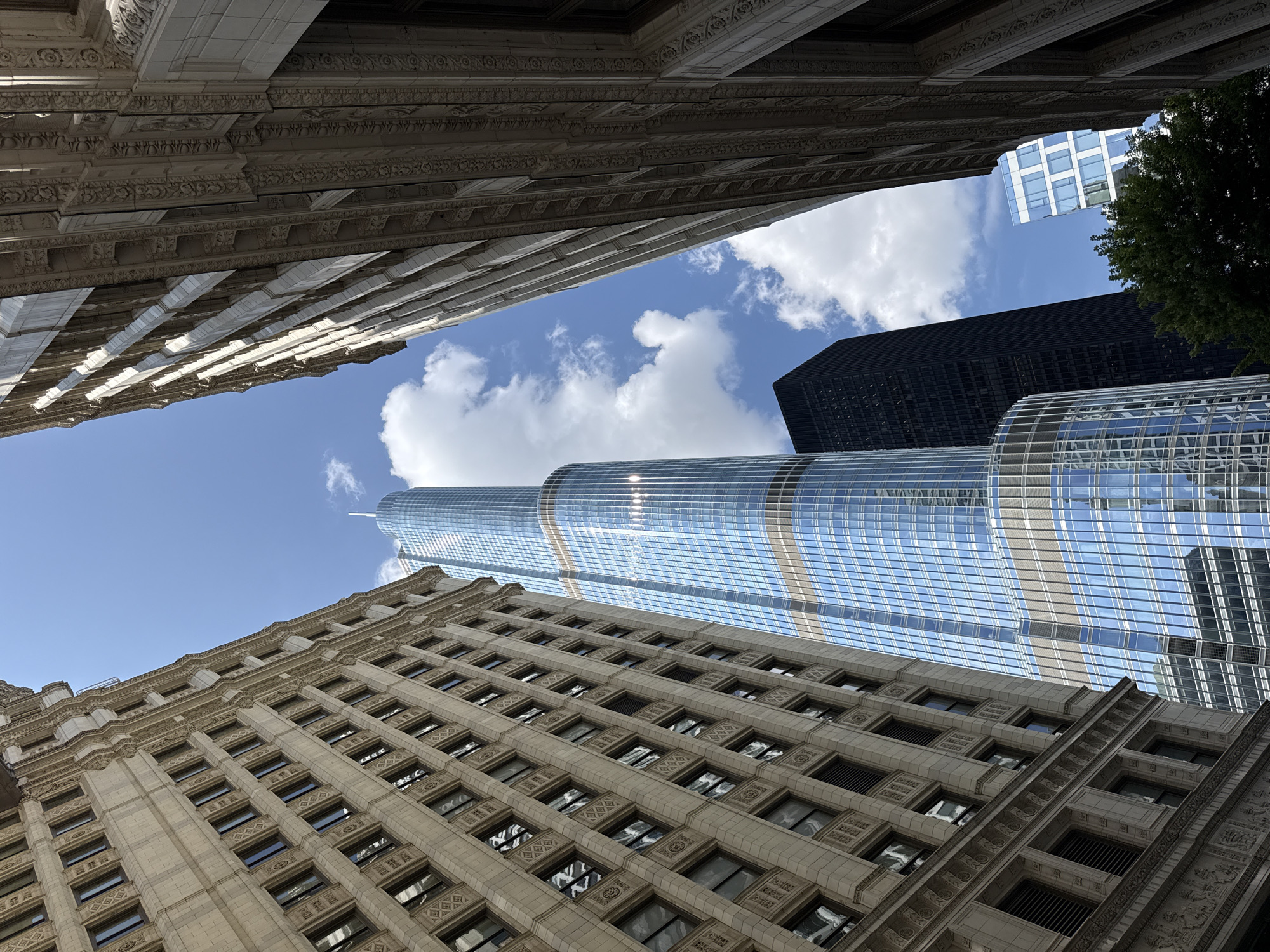 This is a low-angle shot looking up at several skyscrapers against a bright blue sky with scattered white clouds. The foreground is dominated by the detailed facade of an older, ornate building, its stone exterior featuring numerous windows and decorative carvings. Directly above and to the right, a modern skyscraper with a glass facade reflects the sky and surrounding buildings. Further in the background, another dark skyscraper is visible. The image is taken from street level, emphasizing the scale and verticality of the architecture. The sunlight suggests it is daytime, likely midday given the brightness and clear sky. There is no text visible in the image.