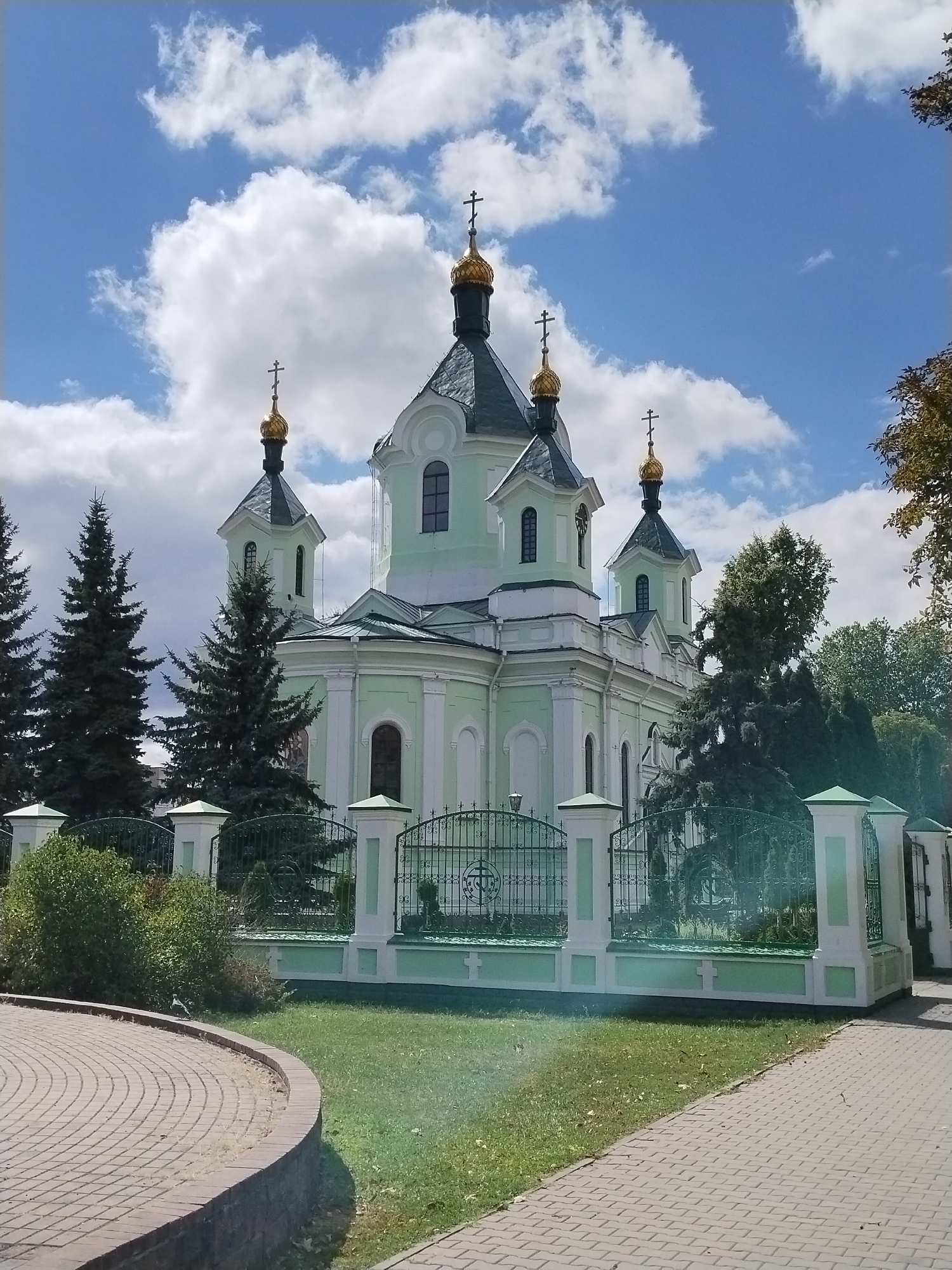 The image depicts the Holy Resurrection Cathedral in Brest, Belarus, on a sunny day with scattered clouds. The light green and white Orthodox church features multiple golden domes topped with crosses. A decorative metal fence with white pillars surrounds the church grounds, separating it from a paved walkway and a grassy area. Several evergreen trees are visible in the background, along with some deciduous trees on the right side of the frame. The foreground shows a curved brick walkway and a patch of grass. No people are visible in the frame.
