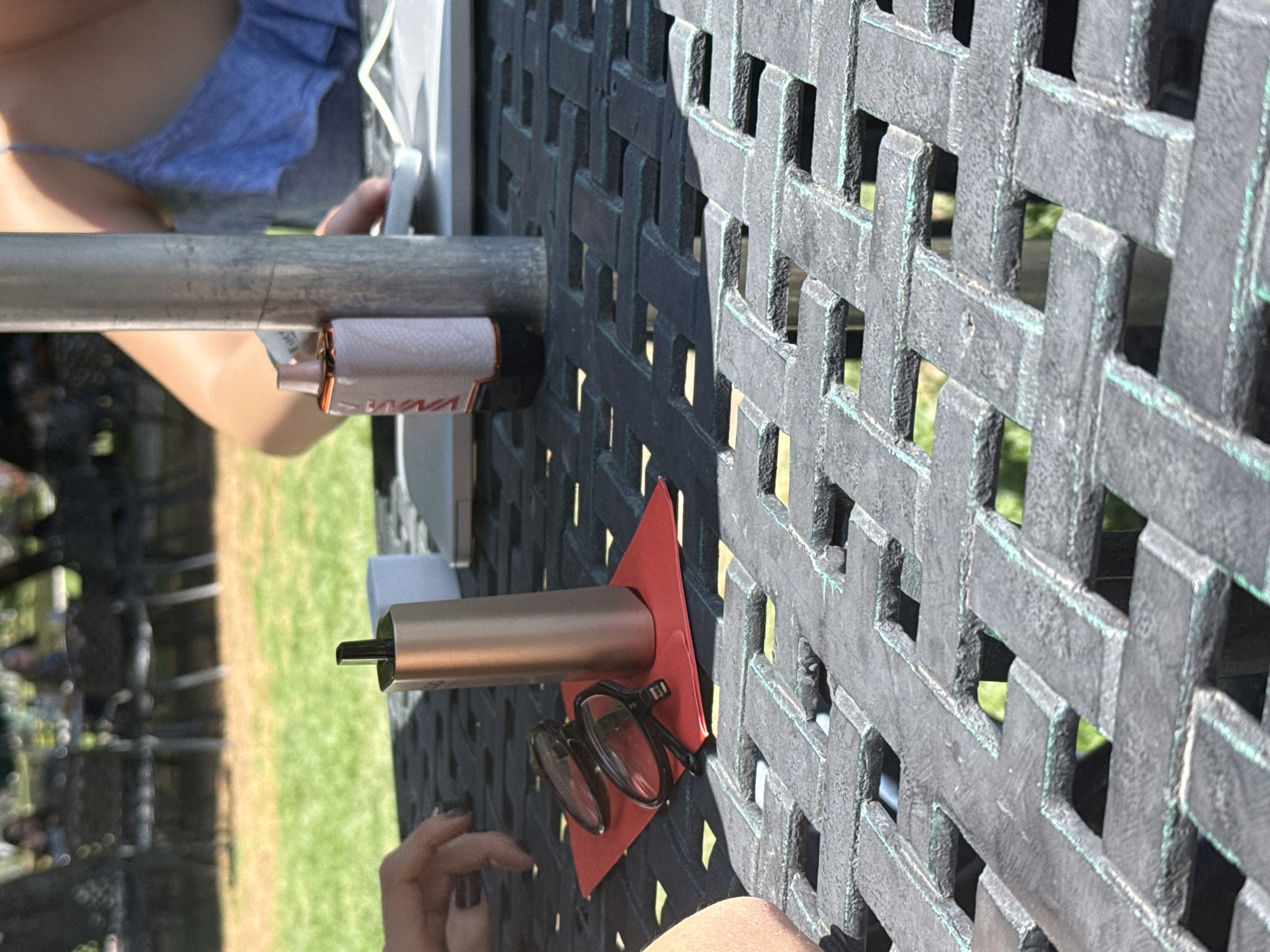 The image shows a black metal patio table with a woven pattern. On the table sits a gold vape pen and black glasses, which rest on a small red square or triangular object. A person is visible on either side of the image. The person on the left is holding a metal pole. The person on the right is wearing a light colored shirt. Outside the table, green grass is visible. The location of San Andrés Cholula, Mexico is not verifiable from the image alone.
