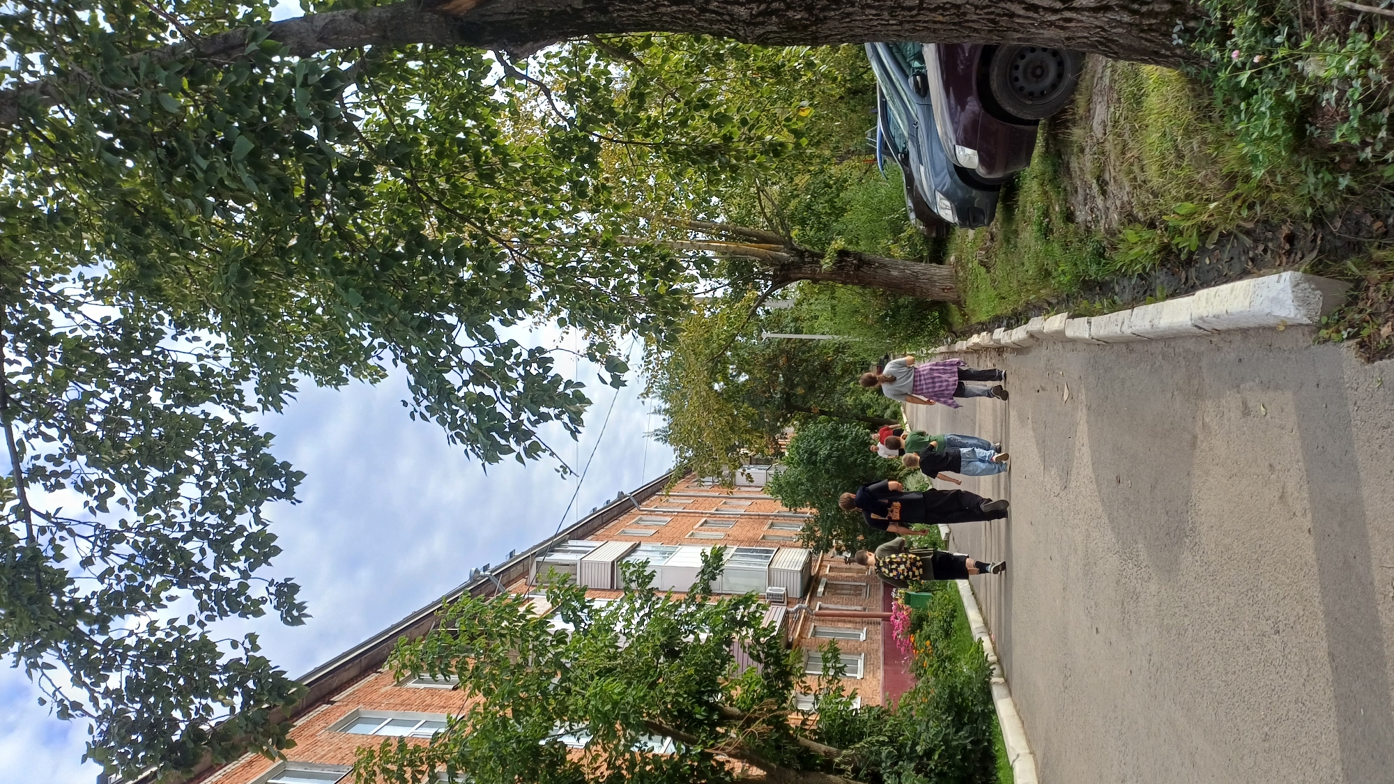 A family of five, two adults and three children, are walking away from the camera on a paved walkway next to a brick apartment building in Izhevsk, Russia. The building is multi-story with balconies and windows. Trees with green leaves line the walkway and the area where several cars are parked. The sky is cloudy.