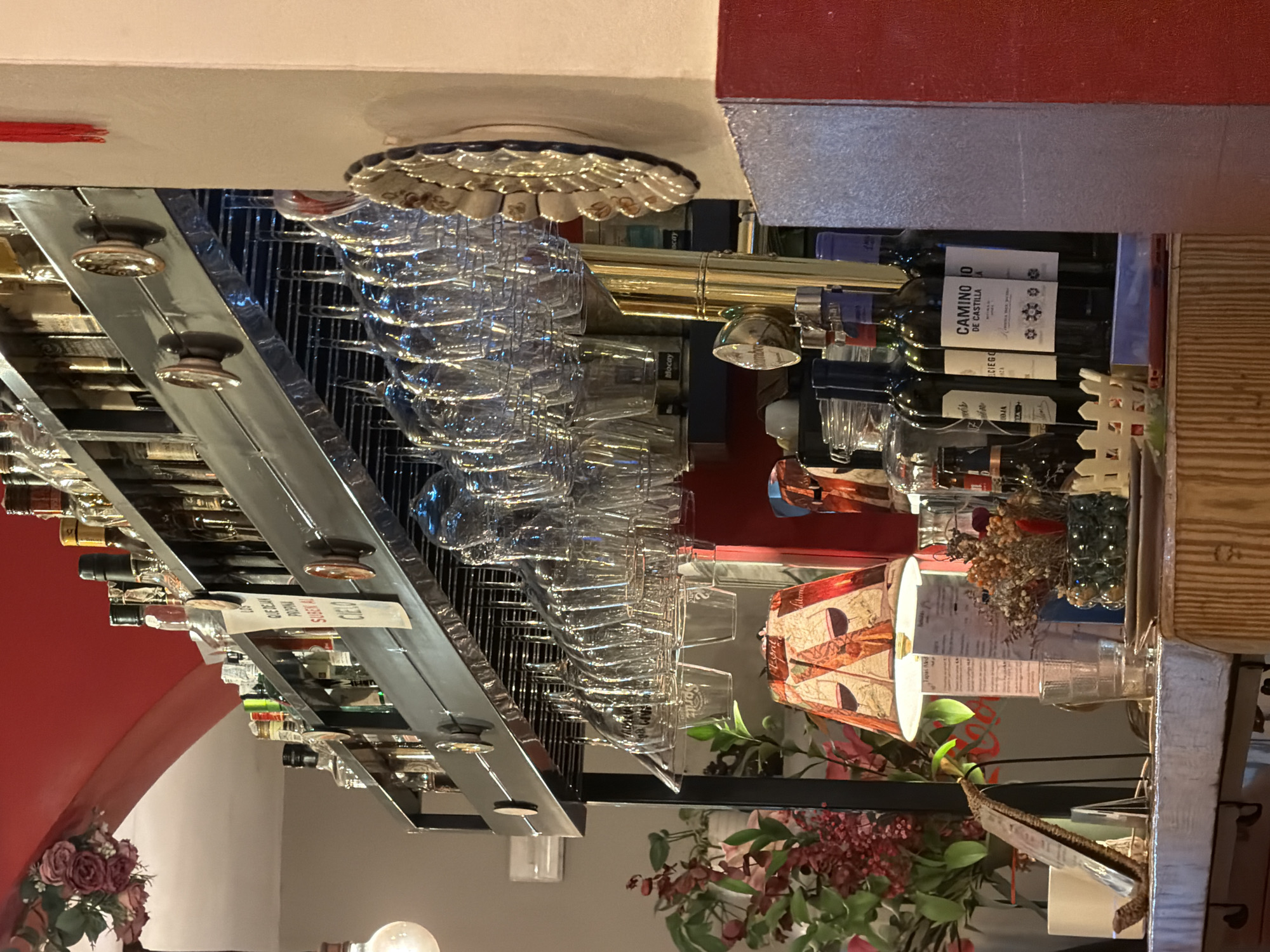 The image shows a bar in Madrid, Spain. The perspective is from below, looking up at the bar's structure. Above the bar, there is a shelf holding rows of wine bottles and glassware. The glasses are hanging upside down in a metal rack. Wine bottles sit on the shelf with labels visible, including "CAMINO DE CASTILLA" and "Mocay." Below the shelf, there's a decorative lamp, flowers, and other bar paraphernalia. Red paint is visible on the ceiling, indicating the establishment's decor. There are price tags attached to some of the bottles. The overall impression is of a well-stocked and decorated bar area.
