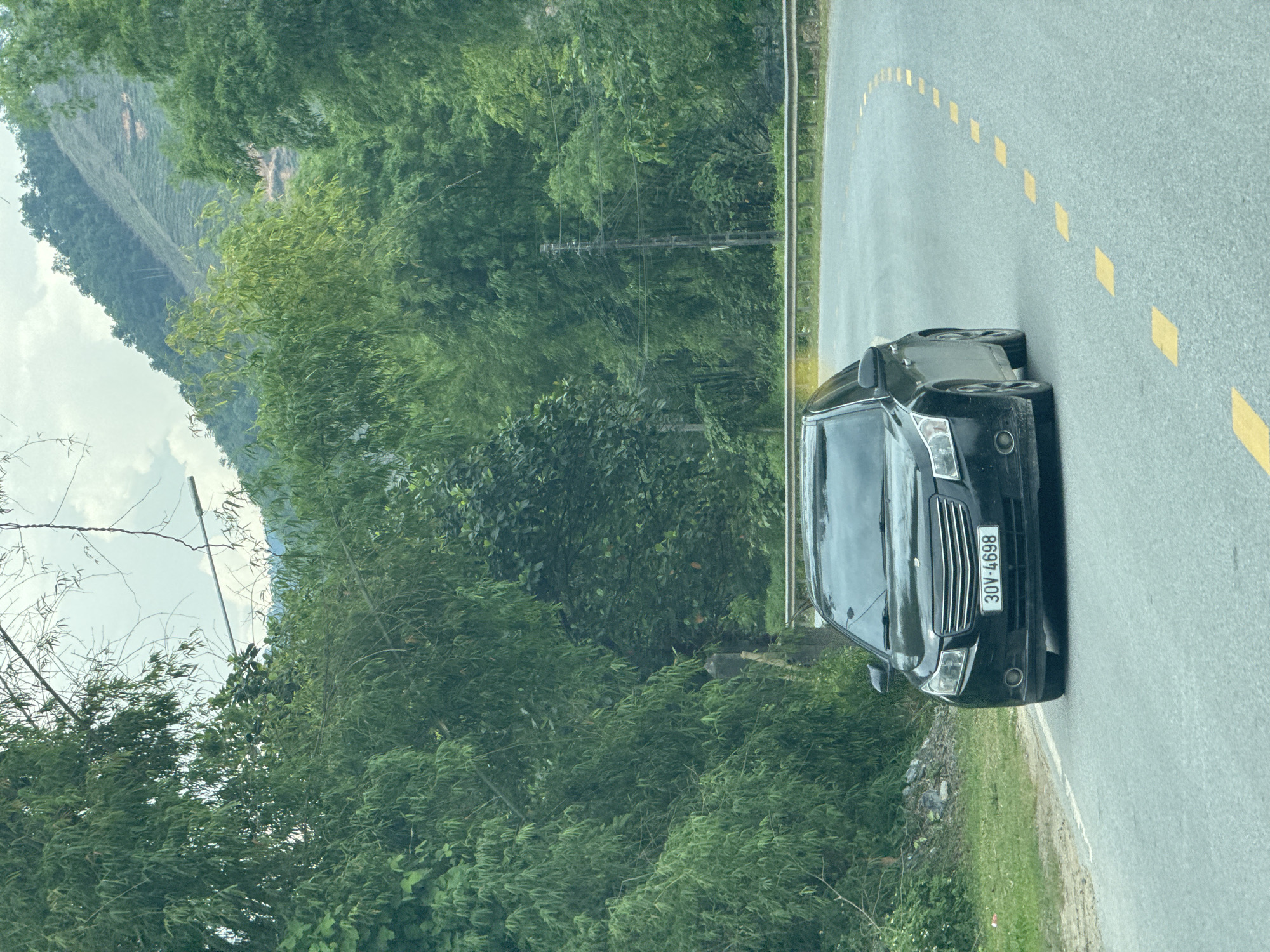 A black sedan, license plate number 30V-4698, is parked on the side of a road marked with dashed yellow lines. The surrounding environment features dense green vegetation, and a distant mountain slope is visible. The scene is located in Hà Giang 2 Ward, Vietnam.