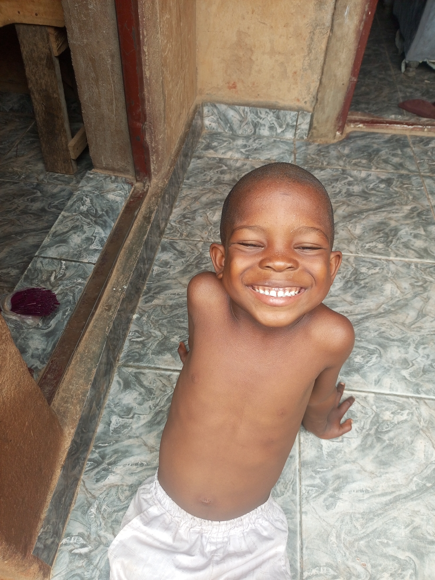 A smiling, shirtless young child wearing white shorts sits on a tiled floor. The location is possibly Tempure, Nigeria, suggested by the subject's appearance. The tiled floor is a light gray with a marbled pattern. The child is positioned near the corner of a wall with a dark wooden frame, potentially a doorway. A wooden structure is visible on the left side of the frame. A small, dark red object sits near the wall. The scene is well-lit, and the focus is on the child's face and smile.