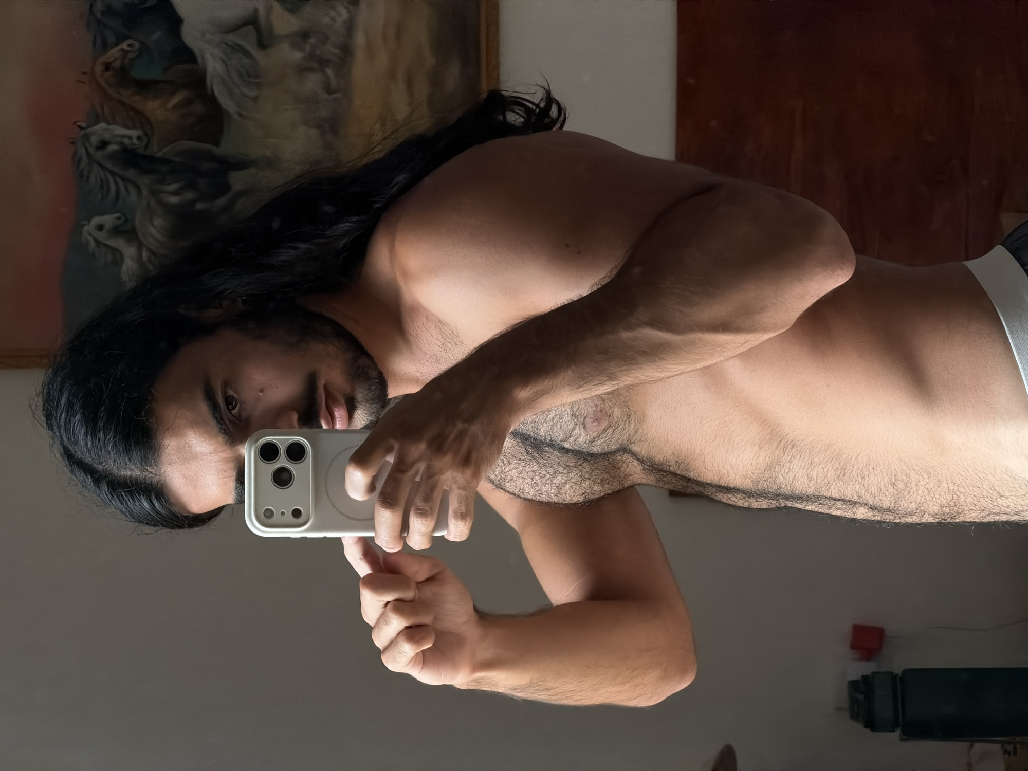 The image displays a man taking a mirror selfie indoors. He has long dark hair and a beard, looking directly into the camera with a neutral expression. The man is shirtless, revealing a muscular torso and chest hair, and the top of his white elastic underwear is visible. He holds a white smartphone in his right hand, featuring multiple camera lenses and a visible circular MagSafe ring on its back, indicating he is taking the photograph himself.
The indoor lighting casts distinct shadows across his body. In the background, a large painting adorns the wall, depicting several horses in motion, rendered in shades of white and darker tones, framed by a wooden border. Below the painting, a small red electrical outlet or switch and a dark, indistinct object are partially visible on the wall. The setting is a private indoor space, likely a room within a residence. The location context for this media file is North Kuta, Indonesia. No discernible text is visible in the image.