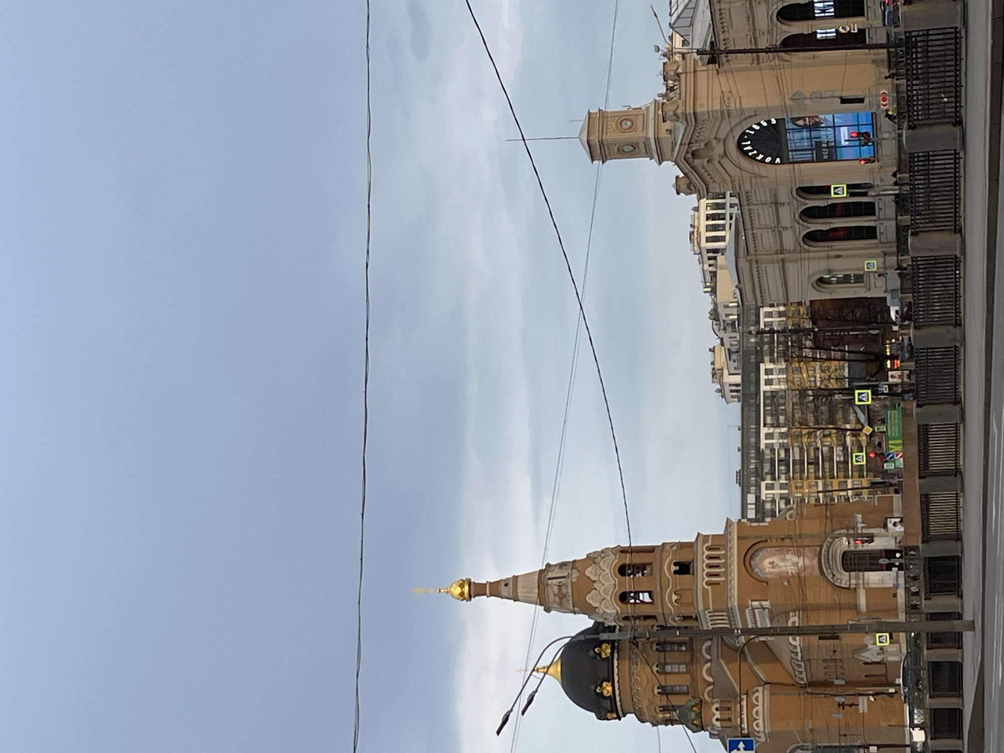 The image depicts a cityscape in Unknown, Unknown, showing two prominent buildings against a cloudy sky. The building in the lower half features a dark dome with a golden spire, surrounded by ornate architectural details in brown and gold. The building in the upper half has a clock tower and an illuminated sign with visible text, as well as traffic lights and crosswalk signs, indicative of a street scene. Power lines traverse the sky. The buildings are rendered in shades of brown and beige, while the sky is a mix of light blues and greys.