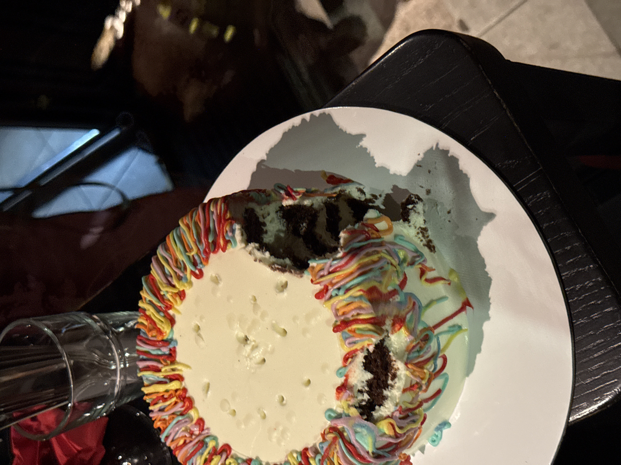 A partially eaten round cake with white frosting and rainbow-colored frosting trim sits on a white plate atop a dark, textured surface, possibly a table. Two slices are missing, exposing a dark cake layer. A glass, seemingly containing straws or sticks, is positioned near the cake. The scene appears to be indoors, given the lighting and surrounding furniture. The location in the city of Belgrade, Serbia, is not determinable from the image. There are no people visible in the image.