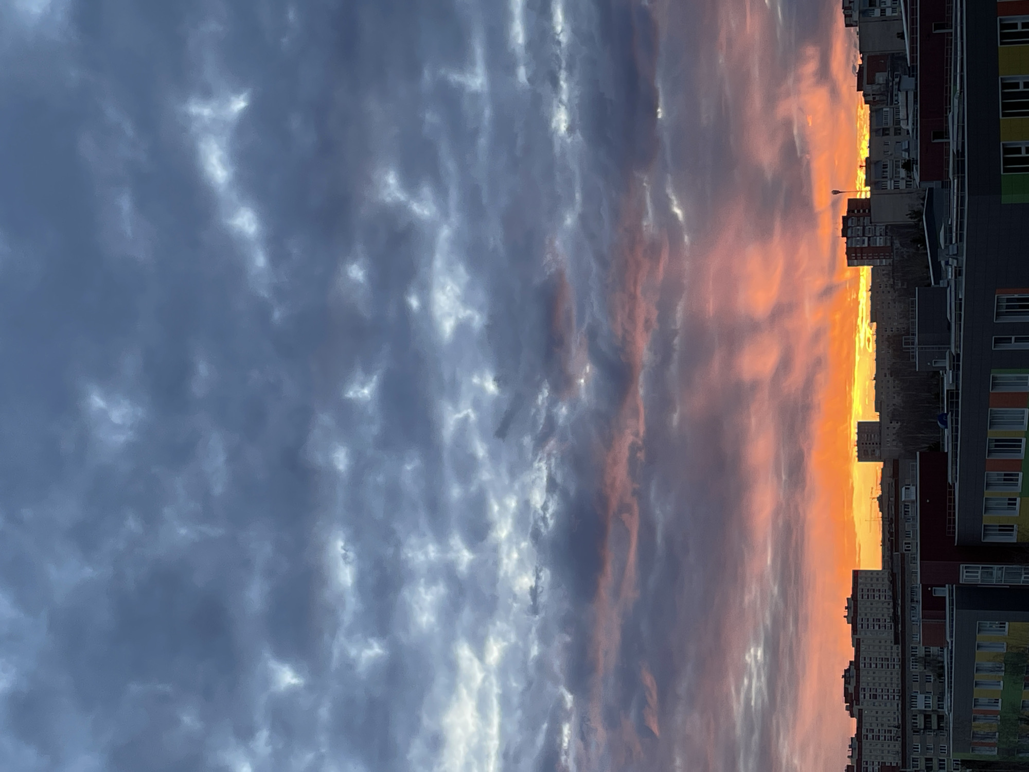 The media file captures an urban landscape at either sunrise or sunset, with a prominent and dramatic sky dominating the scene. The sky, which occupies the upper two-thirds of the image, displays a striking transition of colors. The upper left features dense, dark blue-grey clouds with subtle variations and lighter patches. Moving towards the lower right, the sky explodes into vibrant shades of orange and bright yellow near the horizon, blending into softer pinks and purples higher up, suggesting intense sunlight filtering through or reflecting off the clouds.
Below the colorful horizon, a dense city skyline is visible. Numerous multi-story residential buildings form a dark silhouette against the brightly lit sky. Towards the right side, some buildings are more detailed, revealing multiple windows and facades with distinct colorful panels in yellow, green, orange, and red against a dark grey base. A single lamppost stands silhouetted against the fiery orange sky near the center of the building line. The low angle of the sun, obscured by the buildings, creates a vivid and moody atmosphere. The scene depicts a cloudy but colorful dawn or dusk in an urban setting like Izhevsk, Russia. No discernible text is visible.