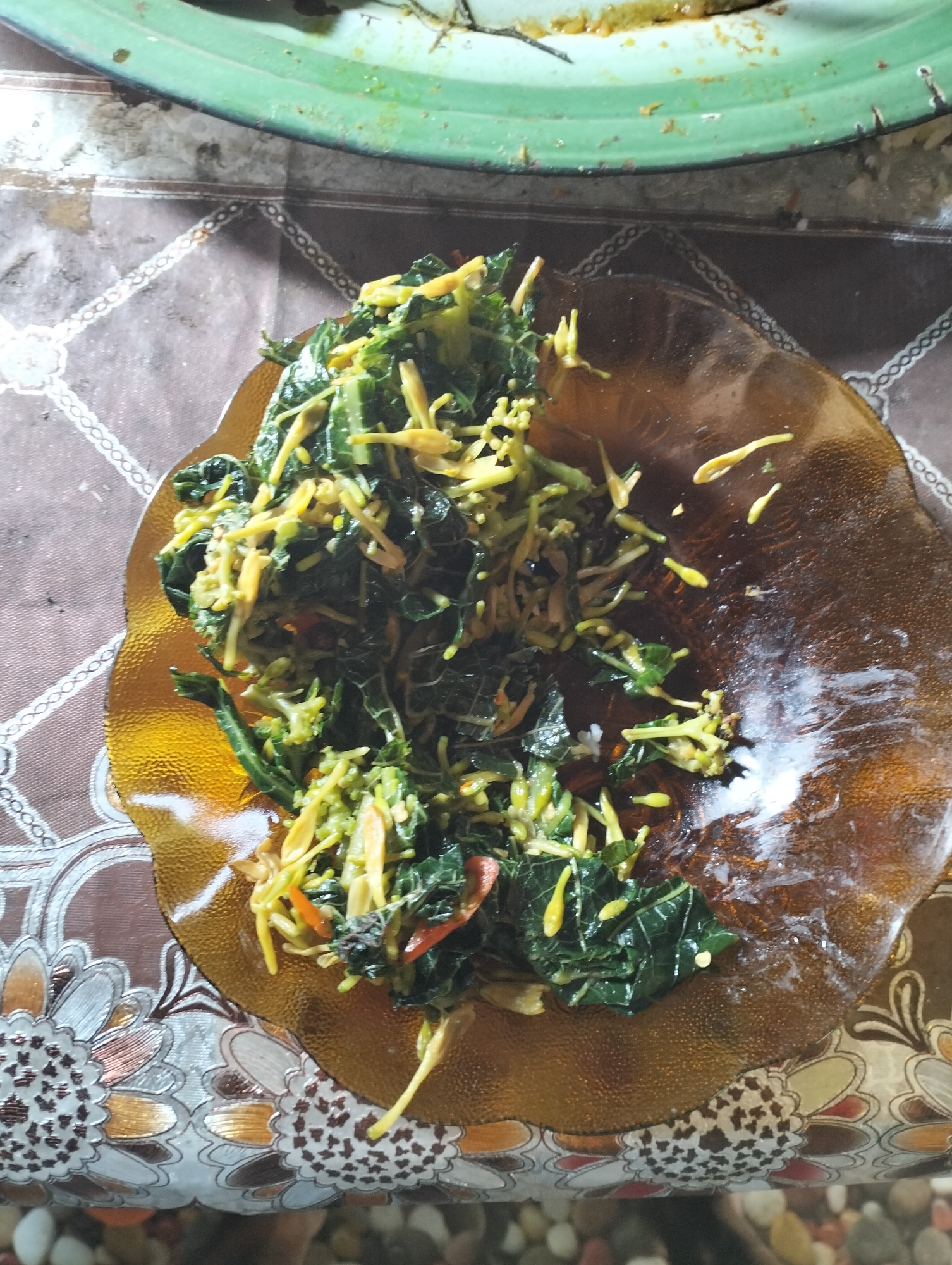 A close-up overhead shot shows a dish of sautéed green vegetables with yellow stalks served in an amber-colored glass plate. The dish is placed on a brown tablecloth with a repeating pattern of white and gold floral and geometric designs. In the background, the edge of a green, shallow bowl or plate is visible, with what appears to be a yellow, semi-liquid food item inside. The lighting is overhead and somewhat harsh, creating reflections on the glass plate and highlighting the textures of the food and tablecloth. The overall scene suggests a meal is being served or has just been prepared in a residential setting, possibly in Mandala Jaya, Indonesia, given the context.