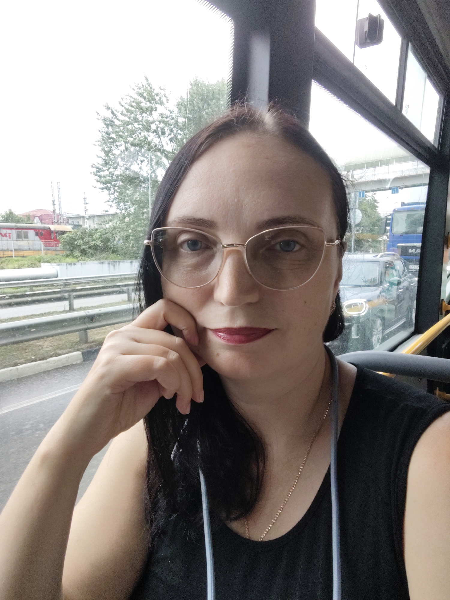 A medium shot shows a woman with dark hair, wearing glasses and a black sleeveless top, sitting inside a vehicle. She is looking at the camera, with her left hand resting on her cheek. The background is blurry, showing a road with cars and trees. The scene appears to be taken during daytime, with overcast skies. The image does not contain any specific landmarks or contextual information to identify the location as Sochi, Russia.