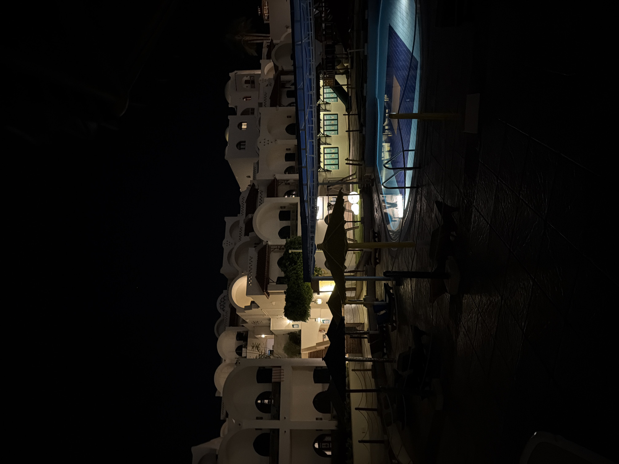 The image depicts a nighttime scene in Sharm El Sheikh, Egypt. On the left side of the image is a multi-story building with white walls and distinctive arched windows and balconies. Adjacent to the building is a swimming pool, illuminated by artificial lighting. Pool furniture, including tables and chairs, are visible in the surrounding area. The overall ambient light is low, making the scene appear dark, with artificial lights providing the main source of illumination. There are no people visible in the scene.