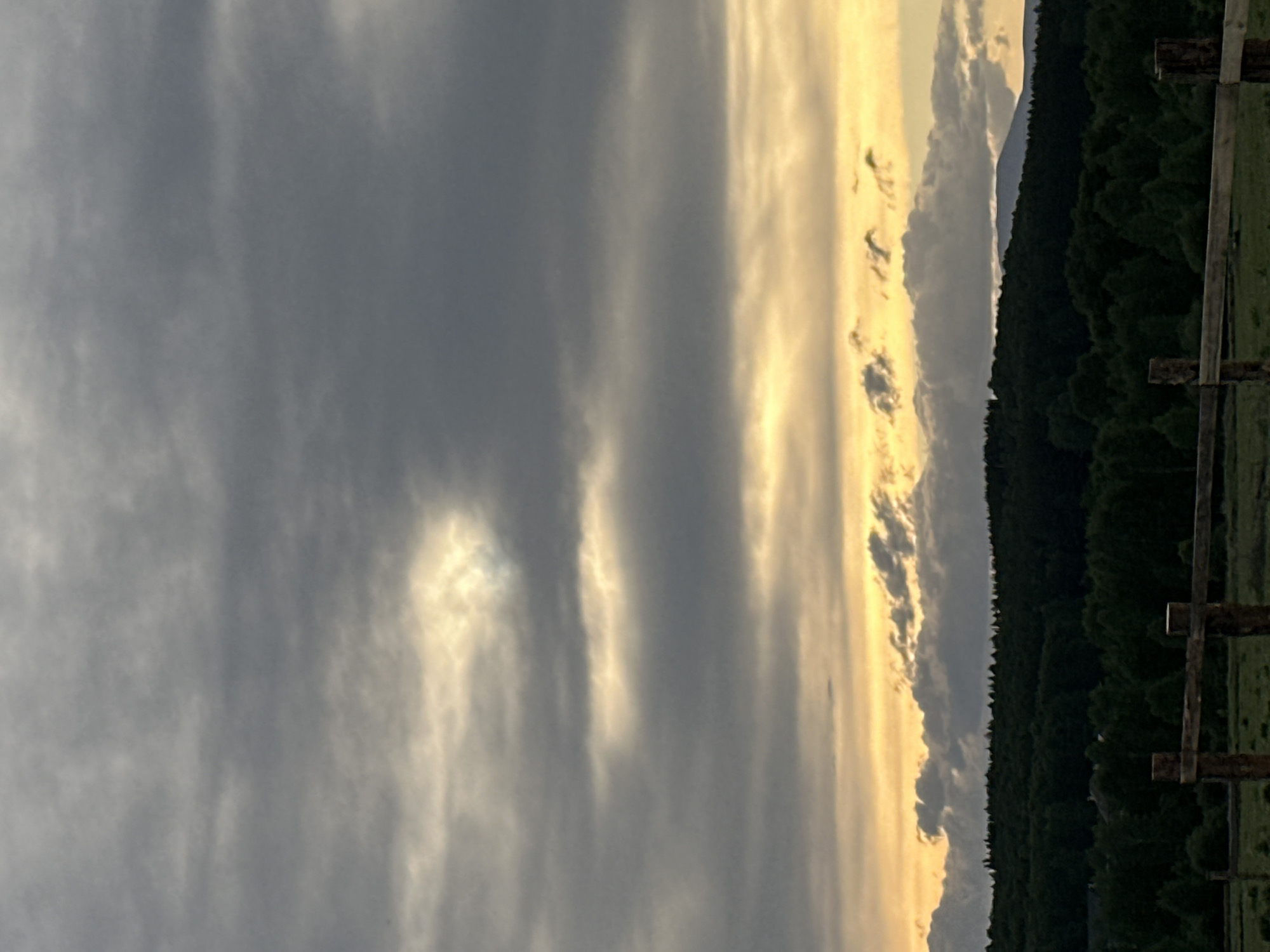 The image depicts a sky with heavy, dark gray clouds occupying a significant portion of the frame. Patches of lighter, golden-toned clouds appear near the horizon, suggesting either sunrise or sunset. Along the right side of the frame, a dense line of green trees forms a boundary between the sky and the ground. A wooden fence with visible posts and rails is positioned in the foreground. In the far distance, a faint outline of a mountain can be seen.