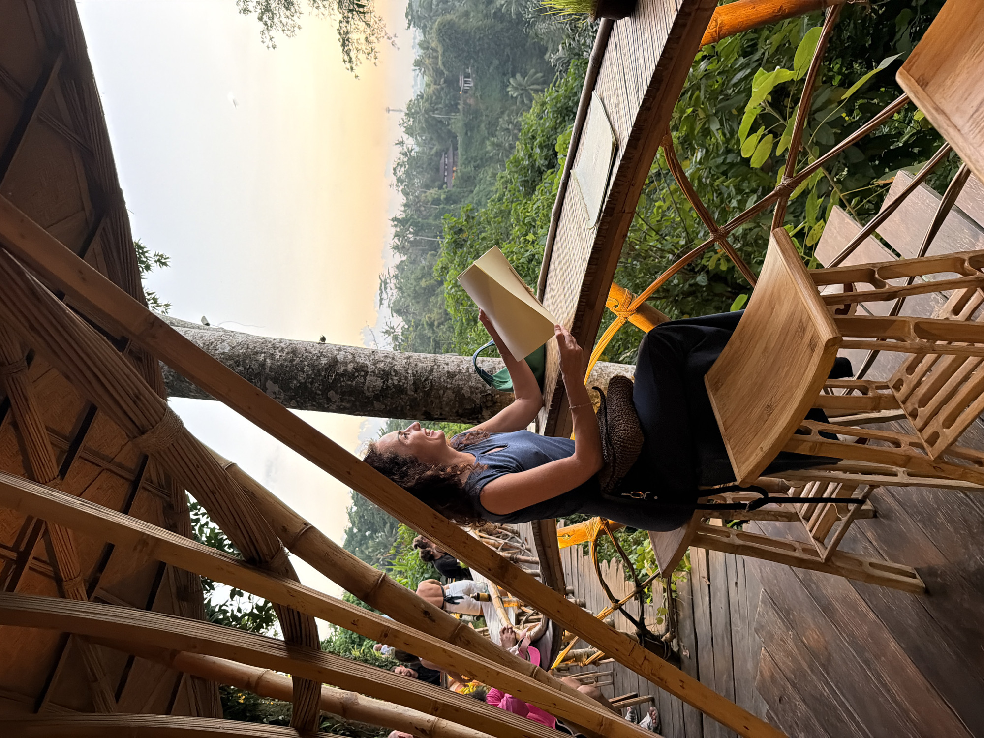 A woman with curly brown hair sits at a bamboo table on a wooden deck overlooking a lush, green valley in Ubud District, Indonesia, at sunset. She is wearing a dark blue sleeveless dress and a woven shoulder bag. In her hands, she holds a pale yellow folder, and she is looking out at the view with a smile. Other patrons are visible in the background, seated at similar tables. The structure is made of bamboo, with a thatched roof. The lighting suggests it is late afternoon or early evening, with the sky displaying warm orange and yellow hues. The scene captures a tranquil moment in a tropical setting.