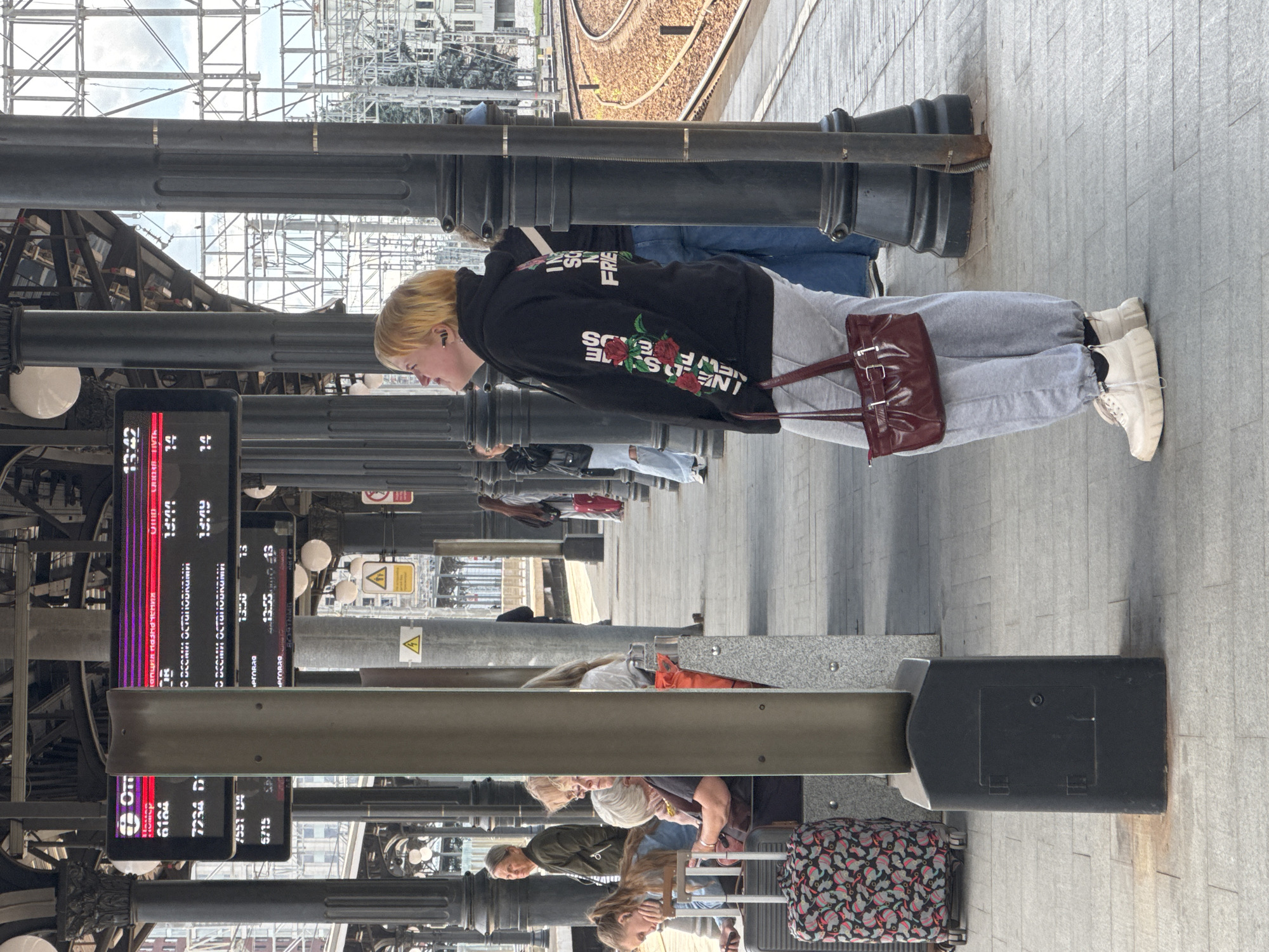 At a Moscow train station, a young woman with short blonde hair stands on a platform, looking up at a digital departure board. She is wearing a black hoodie with rose embroidery on the sleeves and "I NEED SOMEWHERE MY FRIEND" printed on the back, grey sweatpants, and white platform sneakers. She has a maroon shoulder bag. The departure board displays train information, including departure times, destinations, and track numbers in Russian. Several other passengers are visible on the platform, some sitting on benches and others standing. Luggage, including a patterned suitcase, is present. The platform is paved with grey tiles, and large black columns support the station's roof structure. Train tracks and a curved railway line are visible in the background, along with buildings and trees outside the station.