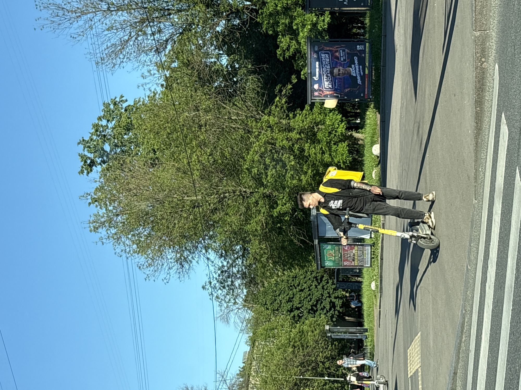 A male individual stands on an asphalt street or sidewalk next to a pedestrian crosswalk under a clear blue sky in Saint Petersburg, Russia. He is oriented slightly to the right of the frame, holding the handlebars of a yellow and grey electric scooter with his right hand. He wears a black top, black pants, and light-colored sneakers, and has a bright yellow square-shaped backpack. Tattoos are visible on his left forearm. His hair is dark with shaved sides and a longer top. Distinct shadows are cast on the asphalt, indicating bright daylight.
In the background, multiple large, leafy green trees dominate the midground. Power lines traverse the upper portion of the sky. To the right of the subject, a large billboard is prominently displayed, advertising "ПЕТРОВСКИЕ АССАМБЛЕИ" (Petrovskie Assamblei or Petrine Assemblies) at "ГАЗПРОМ АРЕНА" (Gazprom Arena) on "30 МАЯ 19:00" (May 30, 19:00), featuring "ВАНЯ ДМИТРИЕНКО" (Vanya Dmitrienko). A smaller, partially visible billboard is to its left. Further in the distance on the left, other people are visible on a pathway, one person clearly seen in a plaid shirt, and another electric scooter is present. The foreground features a white-striped pedestrian crossing.