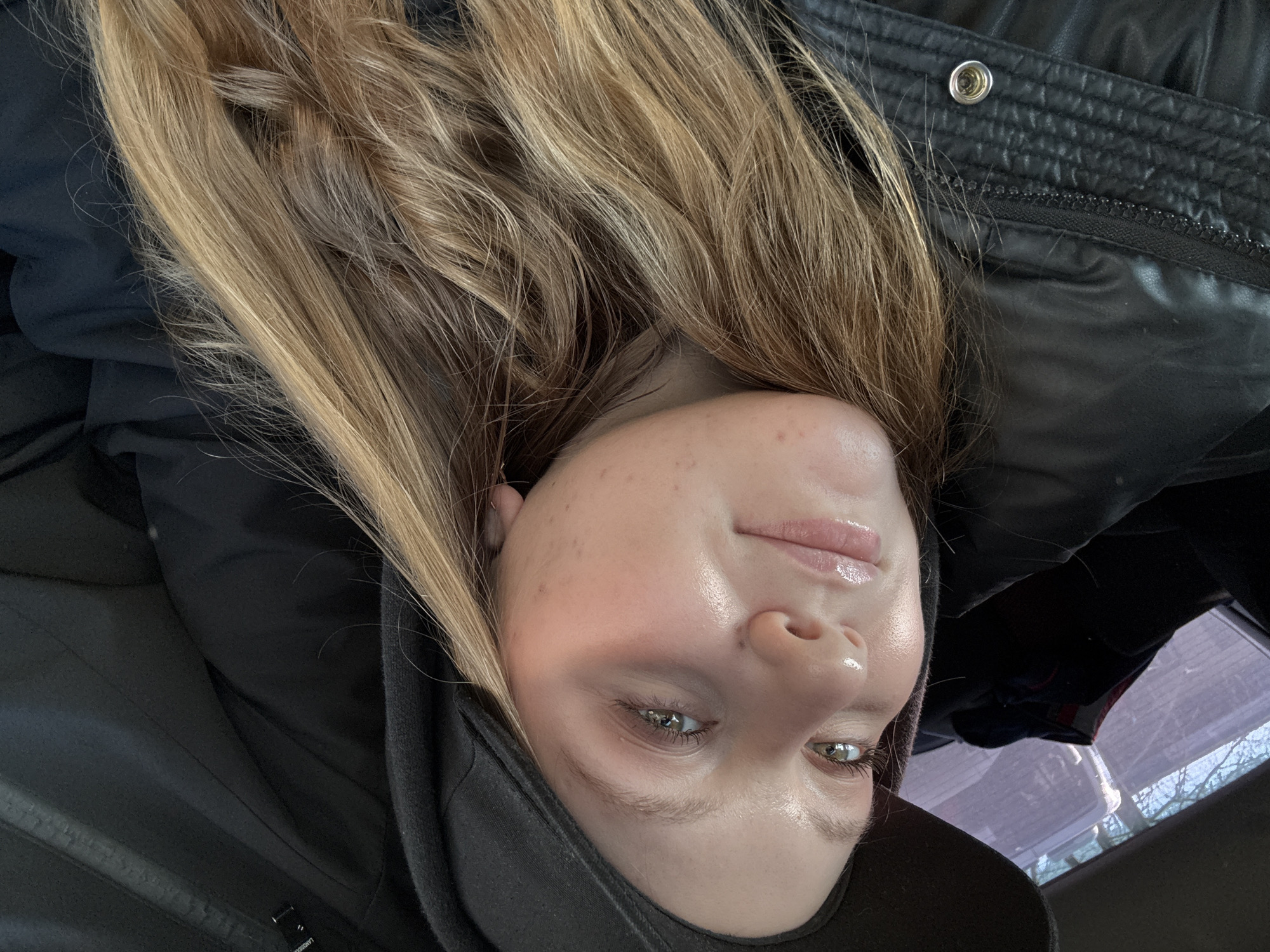 A close-up portrait shows a young adult female, likely in her late teens or early twenties, seated inside a vehicle. Her face, illuminated by natural light, is angled slightly upwards and to the viewer's left, with a gentle expression. She has fair skin, light-colored eyes (appearing greenish-brown), and long, wavy blonde to light brown hair that flows over her shoulders. Her lips appear glossy. She wears a black hooded garment, possibly a hoodie, with the hood pulled up, and a black padded or quilted jacket featuring a visible metallic snap button on the lower left. Through a window visible behind her and to the viewer's right, sparse tree branches against a light blue sky can be observed. The vehicle's interior appears dark, suggesting upholstery and seatbacks. There are no discernible visual cues within the image to confirm the specific location within the city of Chaikovsky, Russia.