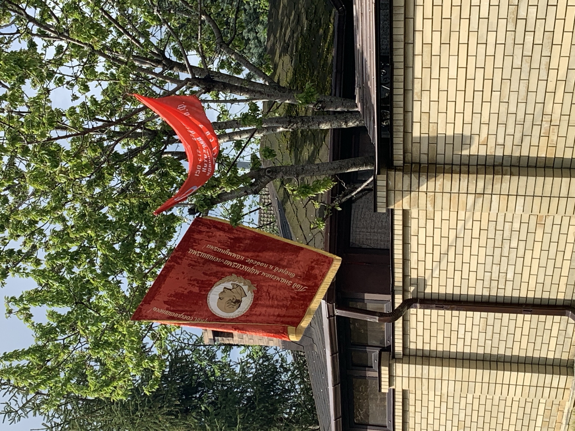 Here is a detailed description of the image:
Two red banners are displayed outdoors. One banner is slightly higher, hanging from the branches of a tree. The other banner is lower, positioned near a building with a yellow brick facade and brown trim. The lower banner has a portrait of Vladimir Lenin on it. Both banners contain text in the Russian language. The background consists of trees and foliage. The location is in the city of Unknown, Unknown.