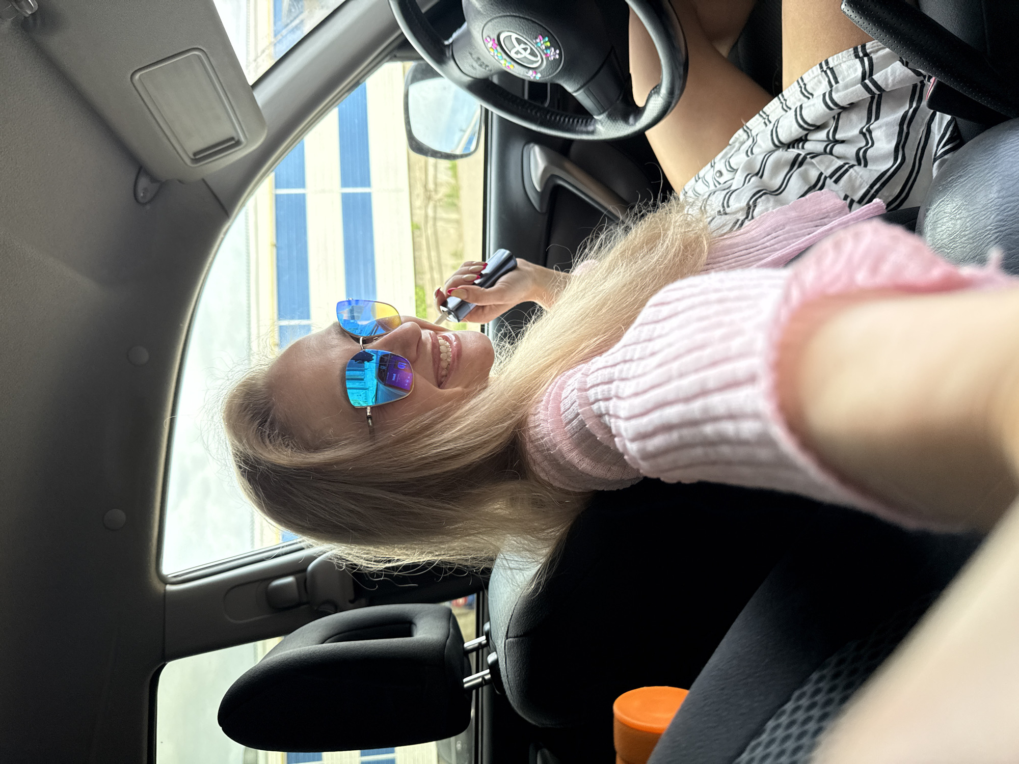 A woman in a pink sweater and striped skirt sits in a car, wearing sunglasses, holding a small black object in her right hand. The car's interior is visible, including the steering wheel with decorative elements, headrest, and dashboard. Outside the car window, there is a building with blue and white vertical stripes. The location is Chaikovsky, Russia.
