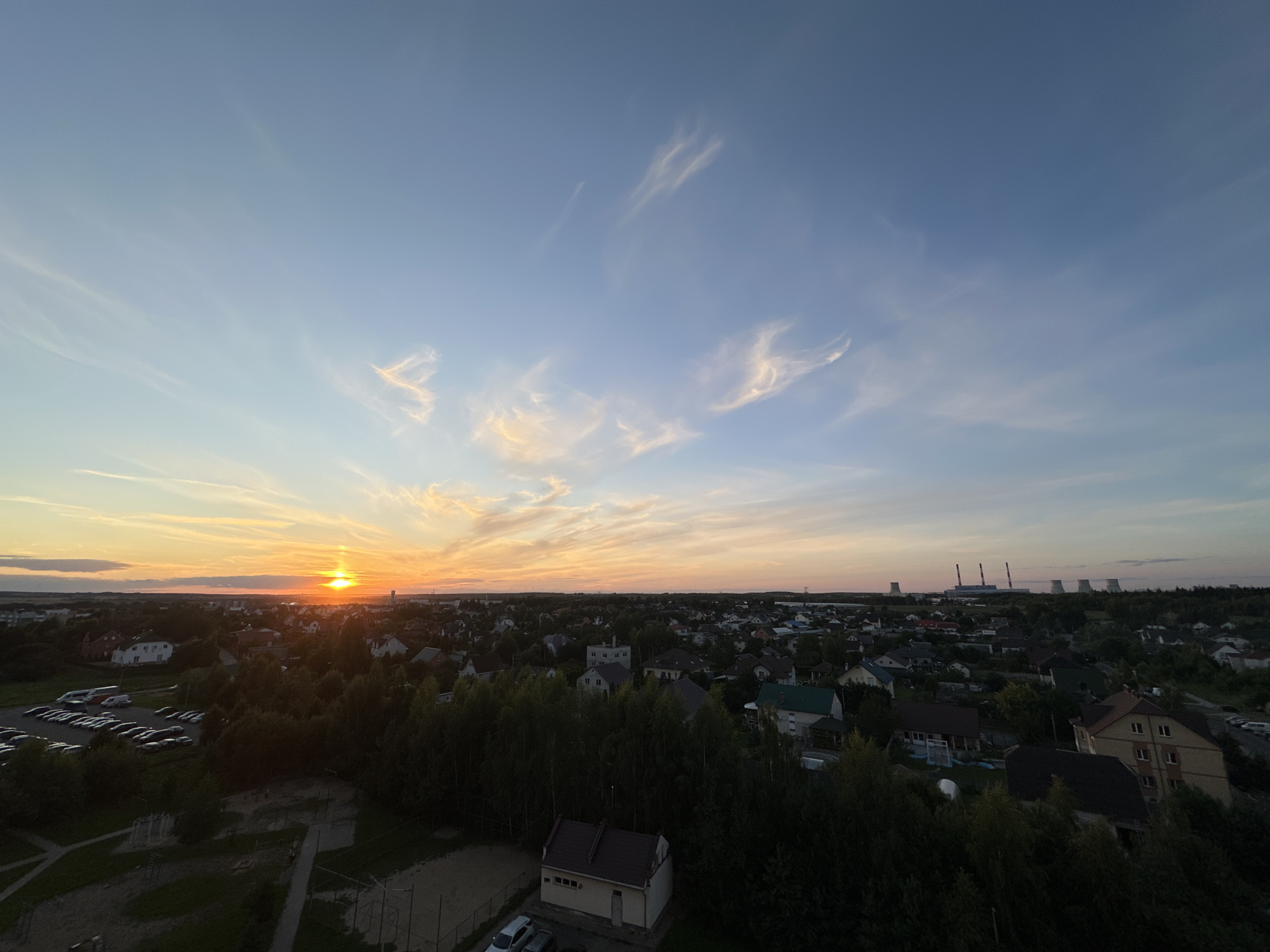 This is an aerial, wide-angle shot of a suburban landscape at sunset, likely taken in or near Шчомысліцкі сельскі Савет, Belarus. The setting sun casts a warm orange glow on the horizon, with scattered wispy clouds tinged with pink and yellow in the bright blue sky. Below, a densely packed residential area with numerous houses, many with pitched roofs, is visible, interspersed with lush green trees that appear to be in full leaf. In the foreground, a section of a park or recreational area is discernible, with playground equipment and sandy patches. To the left, a parking lot is filled with numerous cars. In the distance, several industrial towers, possibly cooling towers of a power plant, punctuate the skyline, silhouetted against the fading light. The overall scene depicts a peaceful, rural-meets-industrial environment during the golden hour.