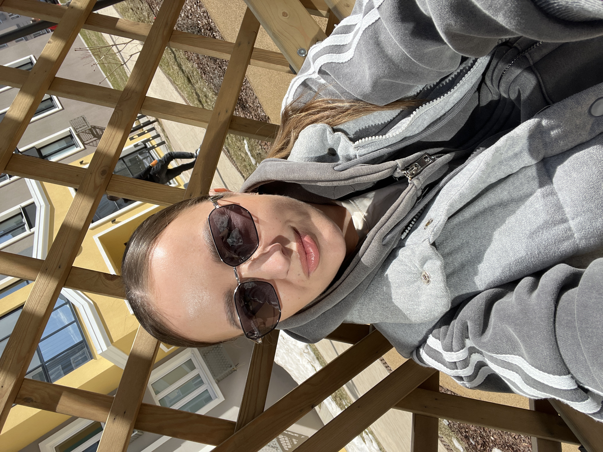 A woman is depicted seated outdoors in a residential or public complex during daylight hours. She is facing the camera with a slight smile, wearing dark, hexagonal-framed sunglasses that reflect the sky and adjacent buildings. Her hair is pulled back. She is dressed in layered grey clothing, including a hooded sweatshirt with the hood up, and an outer grey vest or jacket featuring three white stripes on the left sleeve. She is positioned behind or within a wooden lattice structure, likely a trellis or pergola, which casts strong linear shadows across the scene.
In the immediate background, a building with bright yellow walls and white architectural trim is visible, featuring large rectangular windows reflecting the blue sky. To the right, another building with grey-beige walls is partially visible. A paved pathway runs across the mid-ground, alongside areas of dry grass and patches of melted snow or bare earth. A person wearing dark clothing and light shoes is walking on this pathway, partially obscured by the wooden lattice. Bare tree branches are visible further in the background. The environment suggests an urban outdoor setting during late winter or early spring.