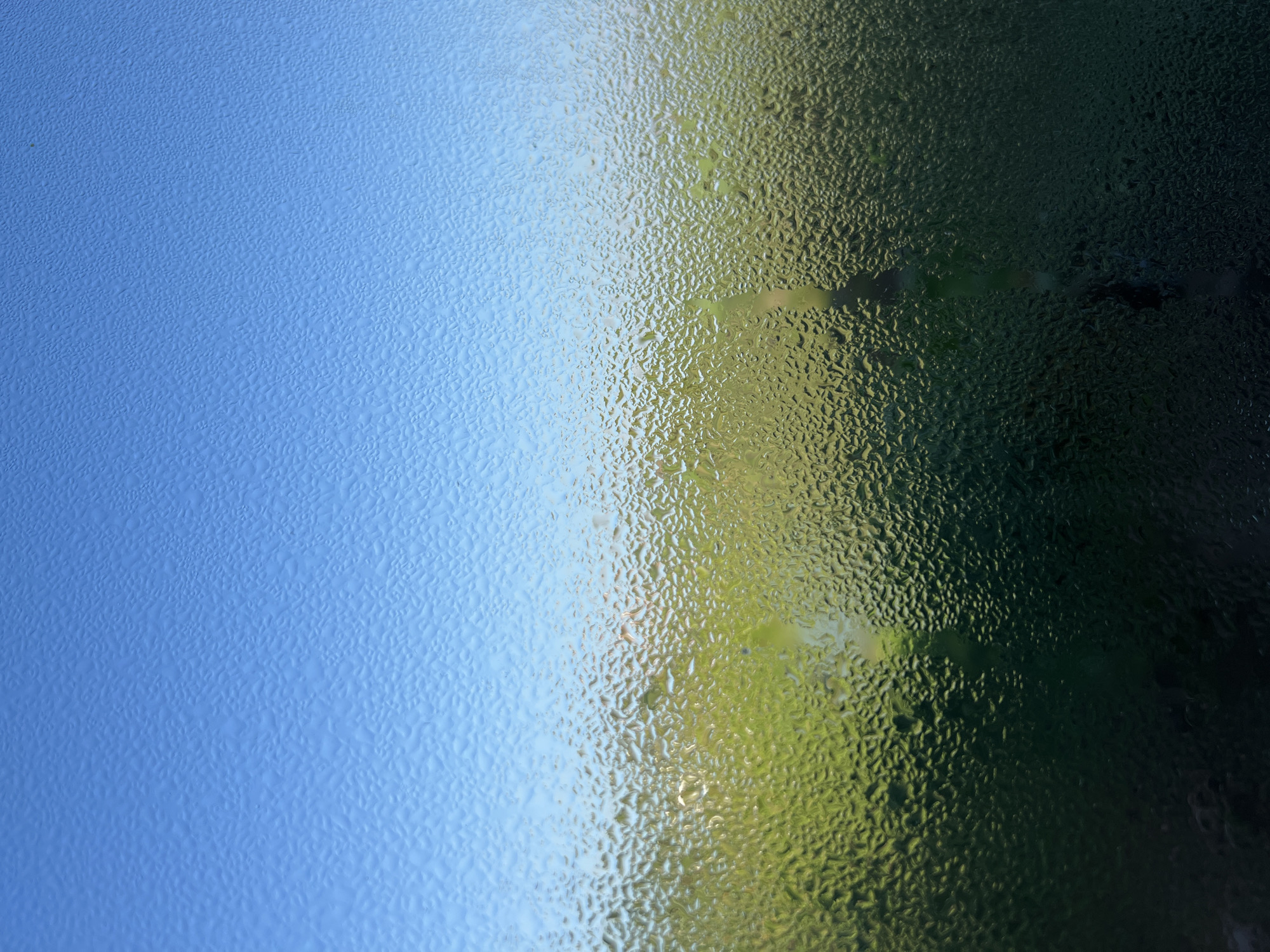 The image is a close-up, textured view of condensation on a windowpane, looking outwards. The upper portion of the image features a clear, bright blue sky, gradually transitioning to a hazy, light blue near the horizon. Below the sky, the window is covered in numerous small water droplets, creating a distorted, shimmering effect.
Through the condensation, a landscape is partially visible. The middle section reveals a bright, undulating expanse of green, likely representing lush vegetation or fields. Hints of lighter colors within this green area might suggest distant structures or vegetation of different hues. The bottom portion of the image is darker, with a deep, shadowed green, indicating a denser or more shaded area of the landscape. The condensation obscures fine details, lending an impressionistic quality to the scene. There are no people, animals, or specific objects clearly identifiable within the frame. The overall impression is one of a natural, possibly rural or suburban, environment viewed through a window on a day with clear skies and atmospheric moisture. The location in North Kuta, Indonesia, suggests a tropical or sub-tropical climate where such condensation might be common due to humidity and temperature differences.