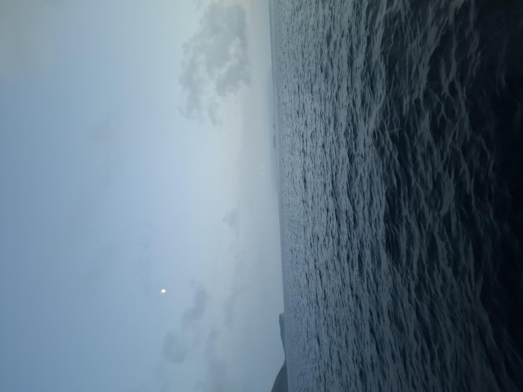 The image depicts a body of water with a distant landmass on the left horizon. The sky is a pale blue with scattered clouds and a crescent moon is visible. The water has a slightly rippled surface, suggesting a gentle breeze. The lighting indicates either early morning or late evening. The scene is serene and vast. Based on the prompt's instruction to include the location of Putyatin, Russia, this image is likely depicting the waters near Putyatin Island, a remote island in the Peter the Great Gulf of the Sea of Japan, off the coast of Primorsky Krai. No people, specific objects, or actions are discernible. The environment is oceanic and atmospheric.