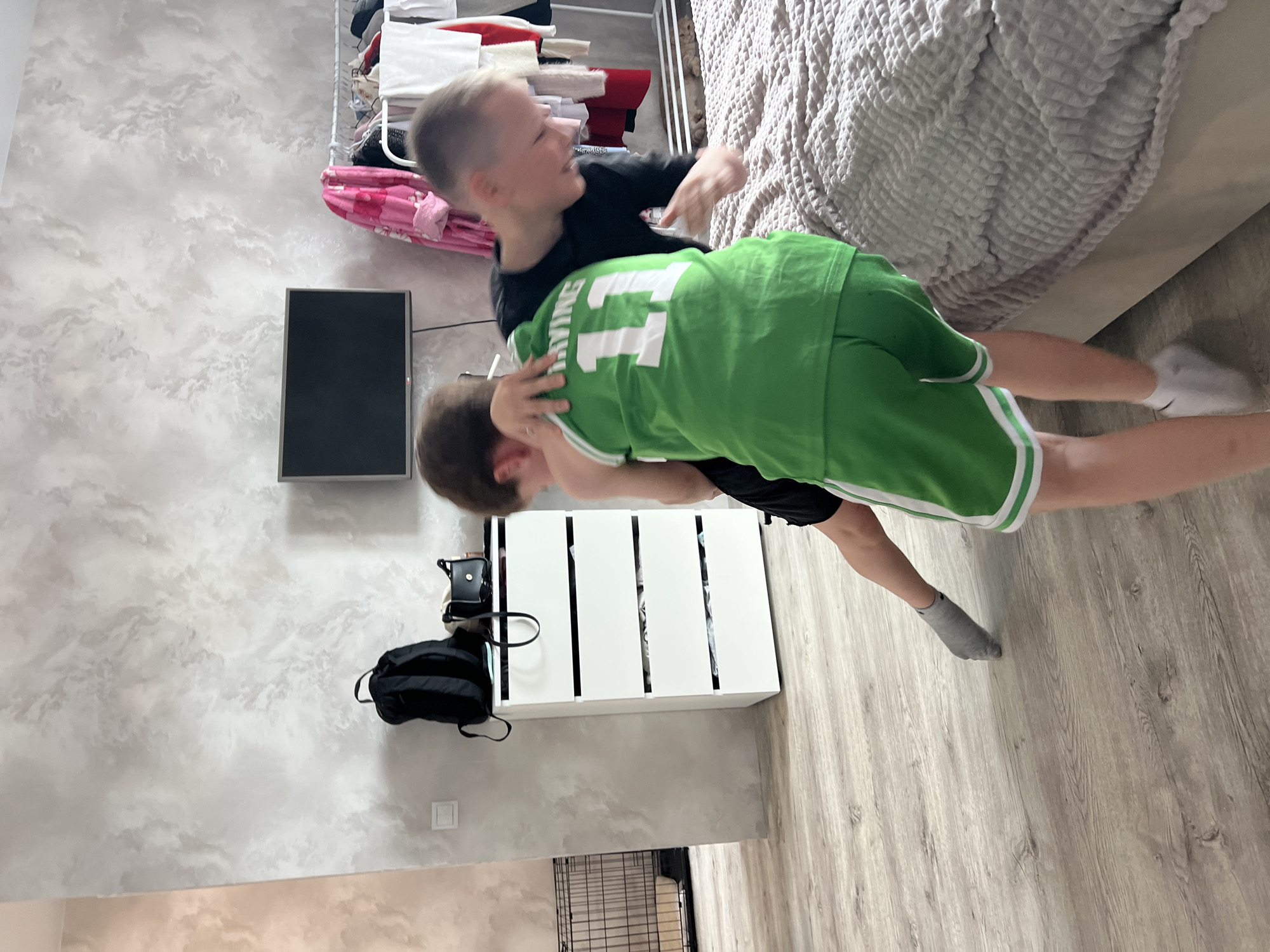 Two boys are wrestling in an indoor setting, possibly a bedroom. One boy wears a black shirt, while the other is clad in a green jersey and shorts with white stripes. The location is suggested to be Izhevsk, Russia, although this cannot be confirmed from the image alone. Visible objects include a bed with a gray cover, a clothing rack with garments, a black bag, a white cabinet with six sections, a television, and a dog cage. The environment appears to be a home interior, with cloud-patterned wallpaper and light-colored wood flooring.