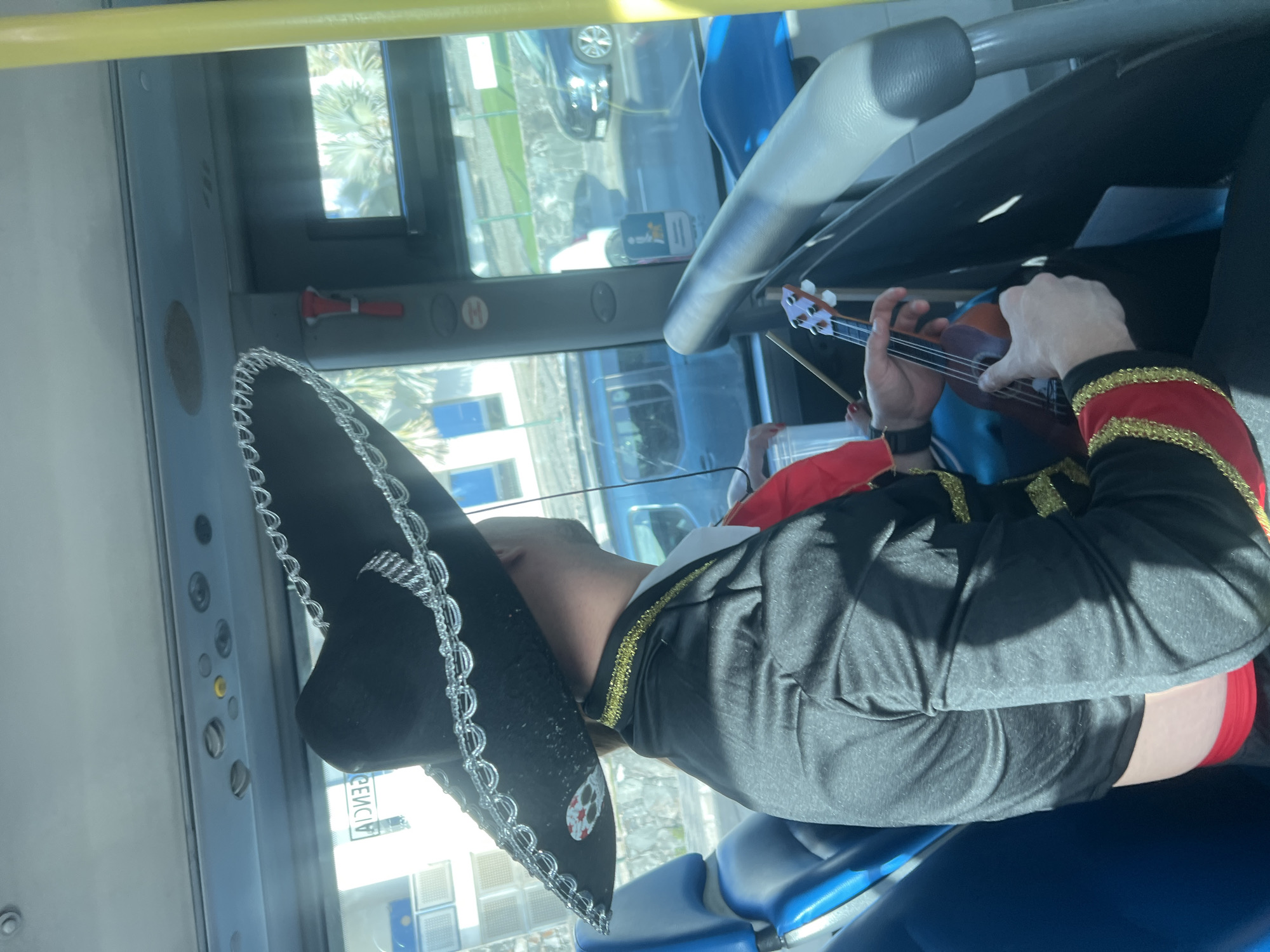 Inside a public bus in San Bartolomé de Tirajana, Spain, a person wearing a black mariachi outfit and a large black sombrero decorated with silver trim is seated. The person is playing a ukulele. Outside the bus windows, buildings and vehicles are visible. The bus interior includes blue plastic seats, a yellow handrail, and various bus fixtures. The light suggests a sunny day.