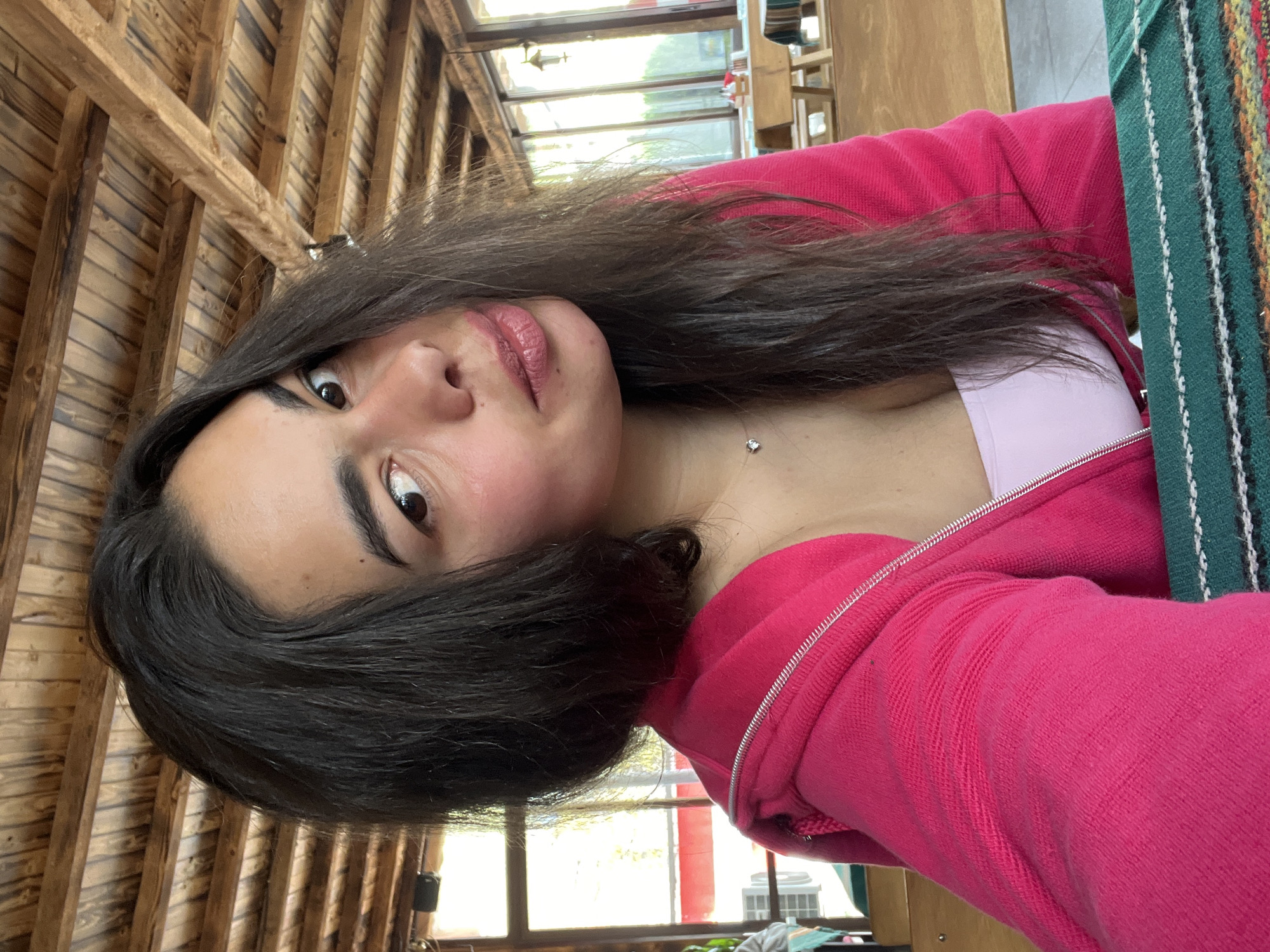 A young woman with dark hair is wearing a pink zip-up hoodie over a light pink top. She is indoors, likely in a building with a wooden ceiling and large windows. There is a green and white patterned fabric object near her. The location is assumed to be Troyan, Bulgaria, though no features confirm this.