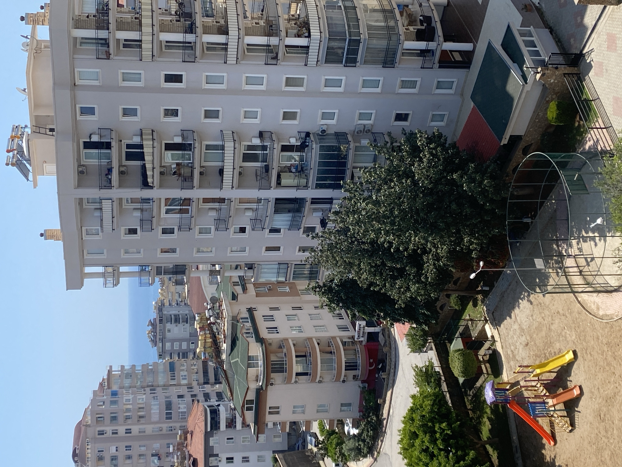 This elevated daytime shot provides a comprehensive view of a residential neighborhood in Alanya, Turkey, under clear, sunny skies. The image is dominated by a tall, modern apartment building on the left, characterized by numerous balconies with dark railings, light grey exterior, and square windows. Many balconies are equipped with air conditioning units, and solar water heaters are visible on the upper floors and roof.
In the middle ground, a cluster of other residential buildings appears, varying in height and style. Notably, one building features curved balconies and a prominent sign displaying "Çalikaya Pansiyon" (Çalikaya Guesthouse) in Turkish, accompanied by a Turkish flag, confirming the geographical context. The distant horizon reveals the calm blue waters of the Mediterranean Sea.
The foreground on the right side of the image features a children's playground set on a sandy surface. It includes a colorful play structure with red, yellow, and orange slides, alongside a climbing frame. A large, skeletal metal archway and a streetlamp are also present in this area. Abundant green trees are interspersed throughout the scene, providing shade and adding a natural element to the urban landscape. Paved paths or roads snake through the neighborhood. No distinct human figures are clearly visible, but the presence of the playground and residences suggests a community setting.