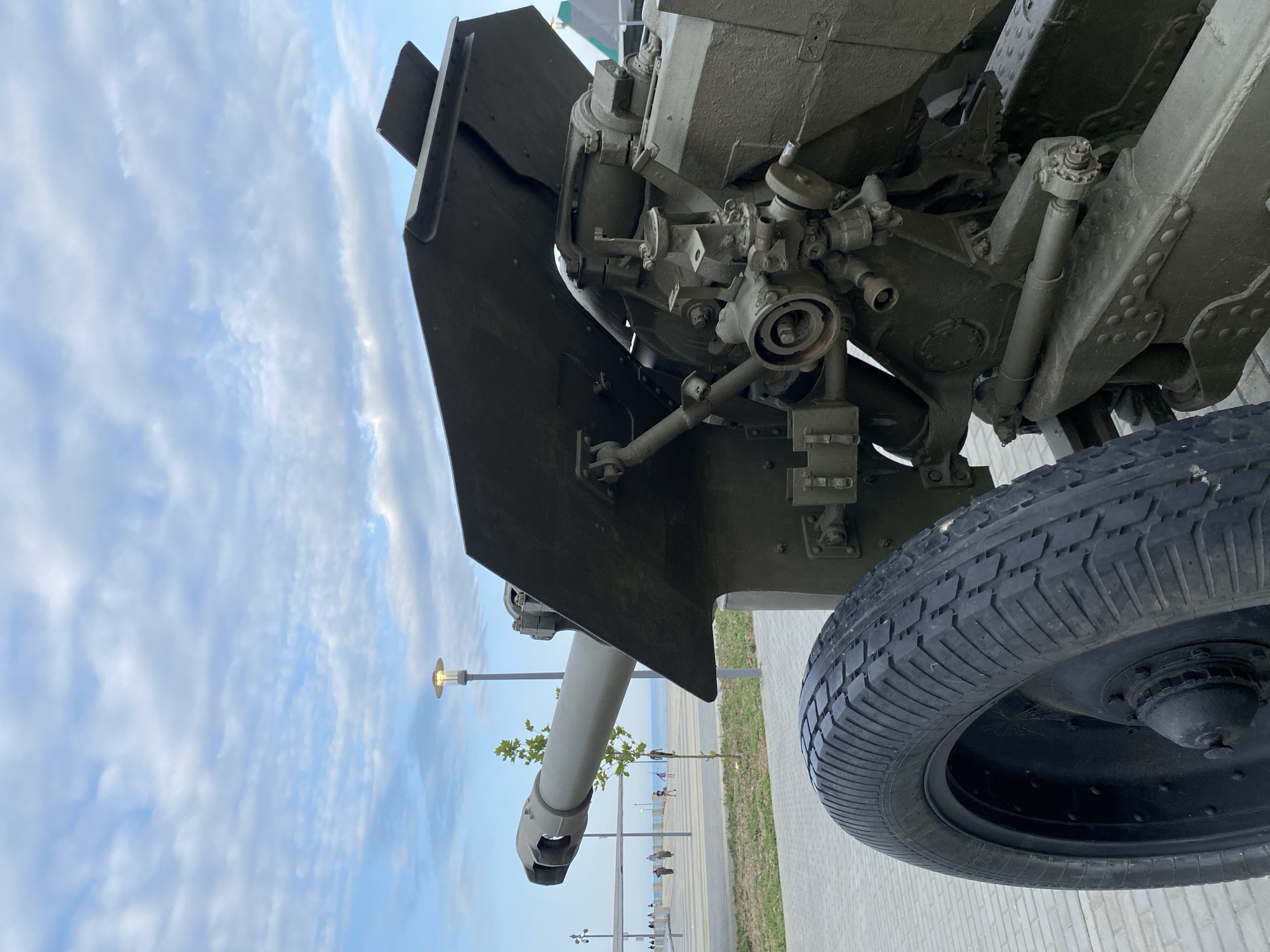 A low-angle, eye-level, medium outdoor shot shows a historical cannon displayed along a paved walkway in Arablyar, Russia. The cannon, painted olive drab, is positioned on the right side of the frame, with its barrel extending to the left. The barrel is thick and appears to be a modern replica or restored artillery piece. A large, black, ridged tire is visible in the foreground on the left. Behind the cannon, a wide, paved walkway stretches towards the sea. Several people are walking along the walkway in the distance, with some appearing to be children. A thin tree stands between the cannon and the walkway. A lamppost with a rounded, illuminated top is visible in the mid-ground, behind the cannon's barrel. The sky above is a pale blue, filled with scattered, wispy white clouds. The overall environment appears to be a park or promenade by the sea.