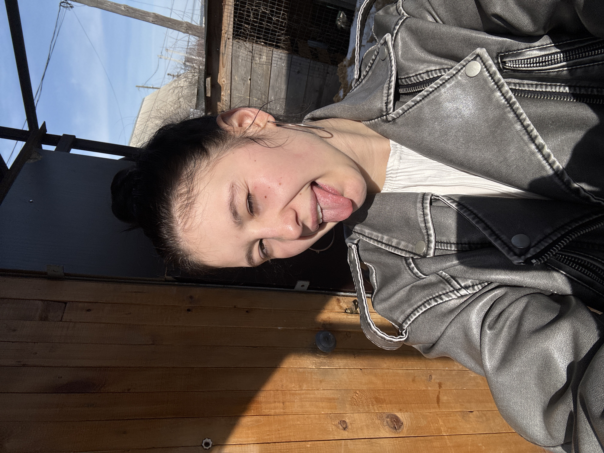 A young woman with dark hair pulled back in a bun is sticking her tongue out. She is wearing a light-colored, possibly white, textured shirt and a gray leather jacket. The background consists of wooden planks and glimpses of a blue sky. Some metal framework is visible above. The location is unclear from the image and can not be specified to be in Фоки, Russia, or any other city.
