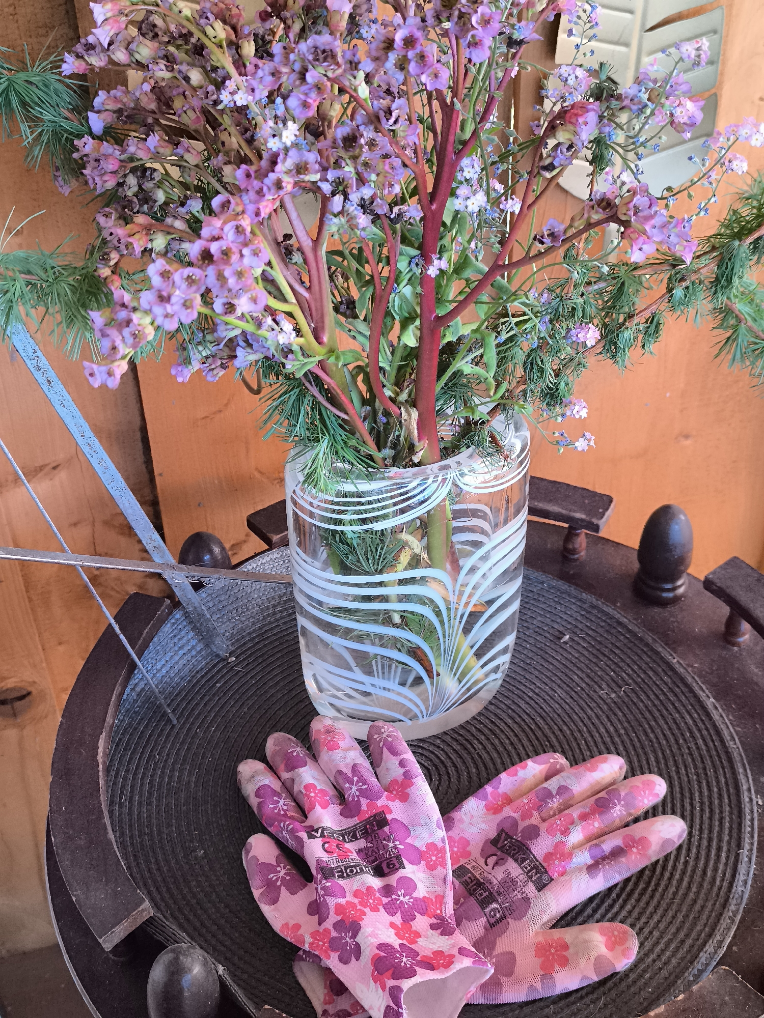A vase of purple flowers and greenery sits atop a dark, circular table. A pair of pink gardening gloves with floral patterns rests on the table's surface, partially obscuring it. A thin, metallic object leans against the table near the vase. The background is a wooden wall. The location is Unknown, Unknown.