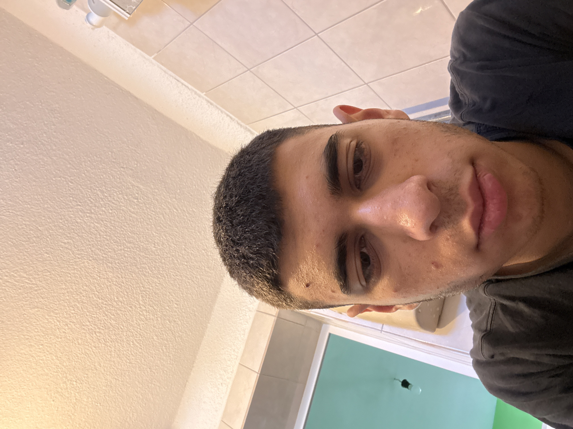 A close-up, indoor selfie of a young man with dark hair, brown eyes, and visible facial acne. He is wearing a dark-colored shirt. The background includes a white tiled bathroom wall, a portion of a mirror, and a green wall, possibly of a shower. There is nothing in the image to indicate the location as Sofia, Bulgaria.